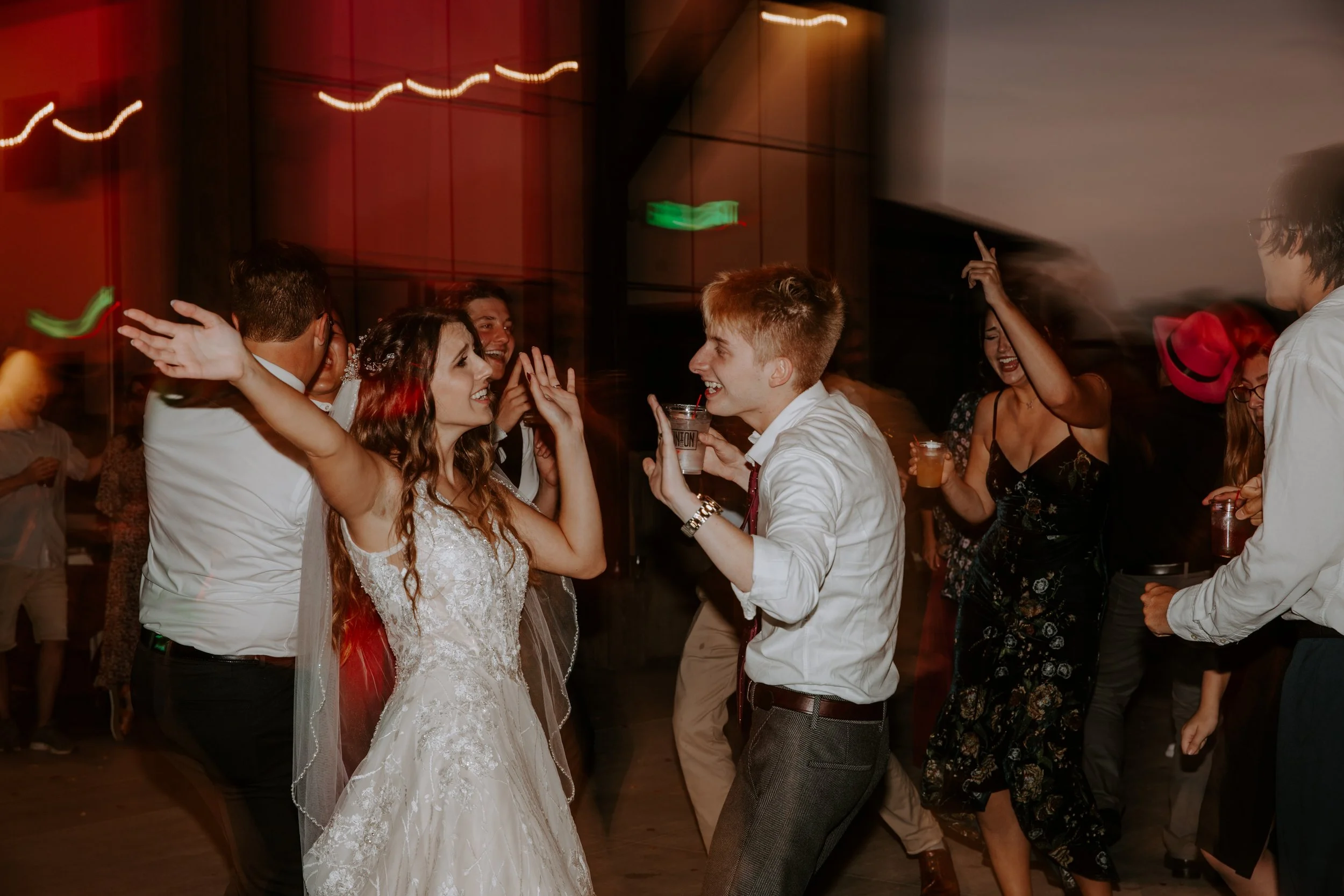 Bride and groom dance together with joyful expressions, surrounded by guests under dim lighting at their reception.