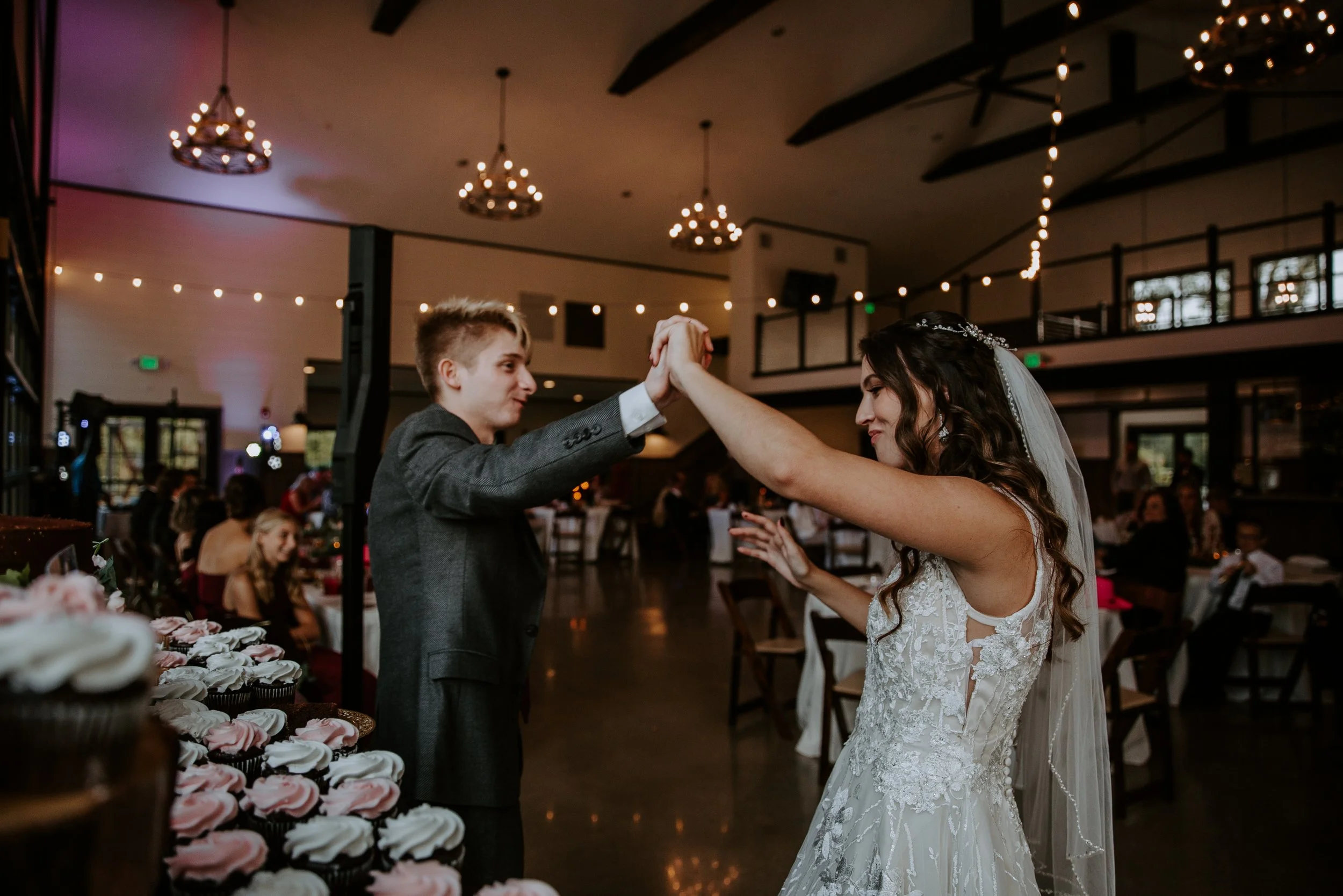 Bride and groom share a dance at their wedding reception, surrounded by tables and a display of cupcakes.