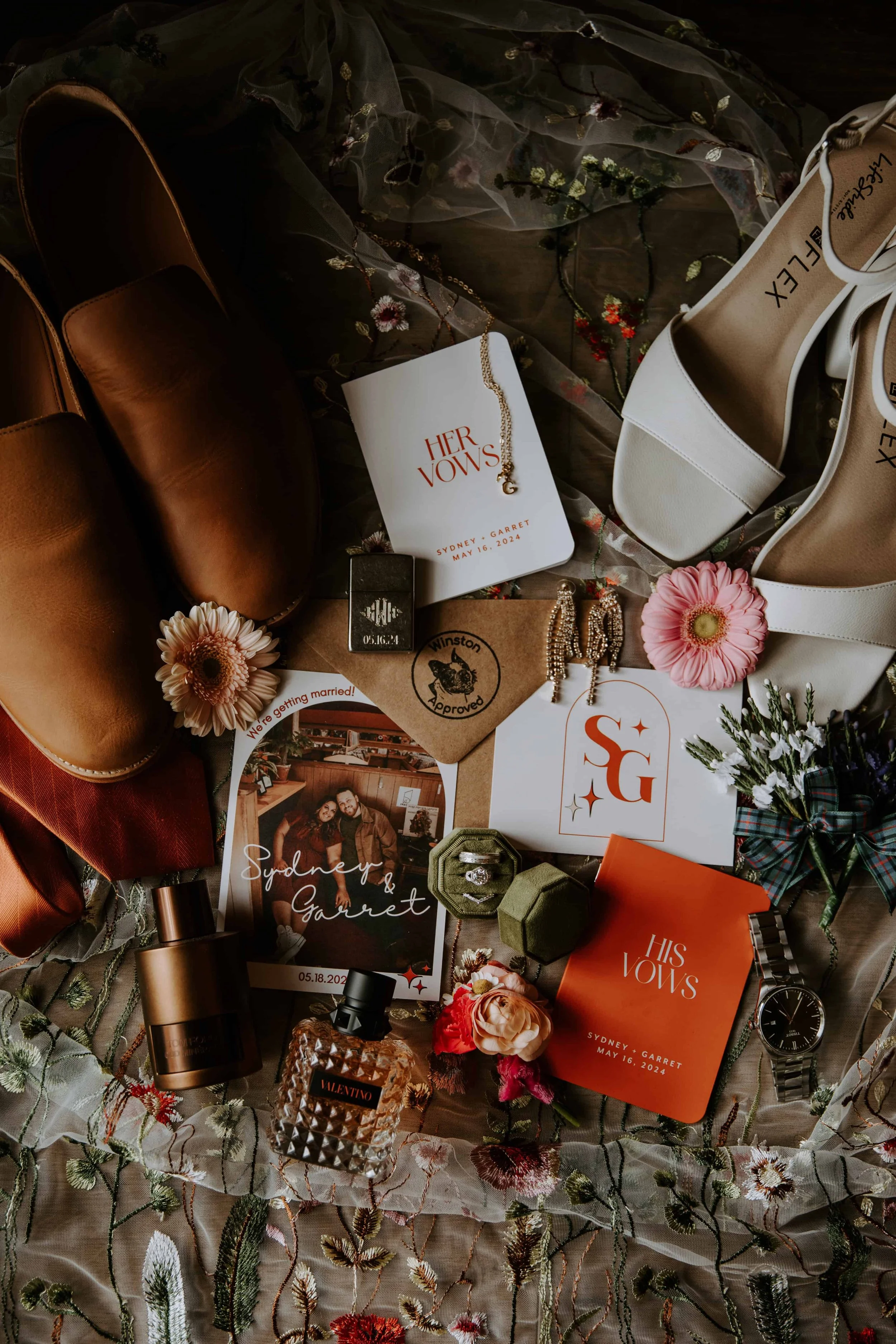 A flat lay of all the wedding details including the bride and groom's shoes, rings, invitation suite, perfume, flowers, and jewelry.