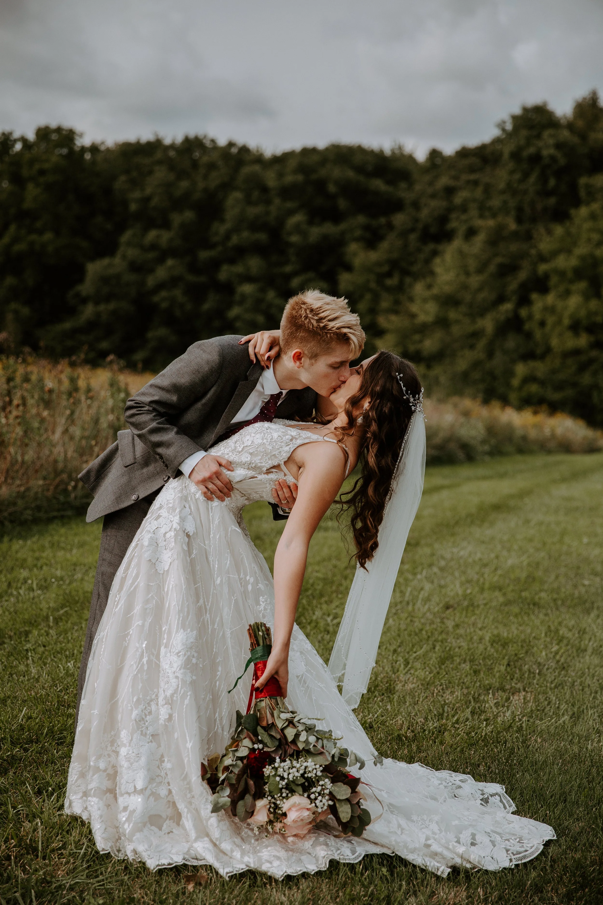 Groom dips the bride for a kiss in a grassy field, as she holds her bouquet and they both smile.