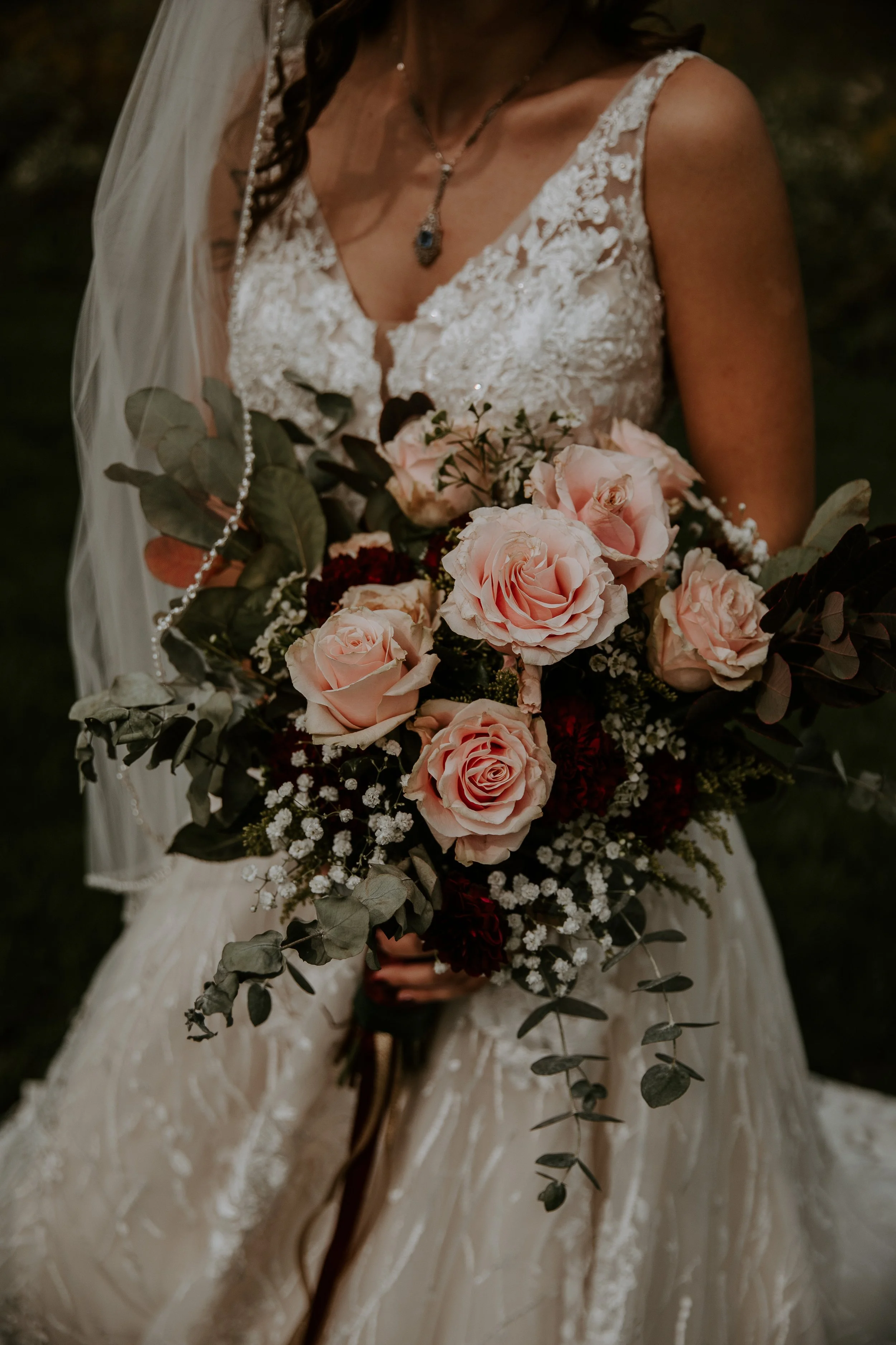 Close-up of the bride holding a bouquet of pink roses and greenery, with the intricate details of her lace wedding dress visible.
