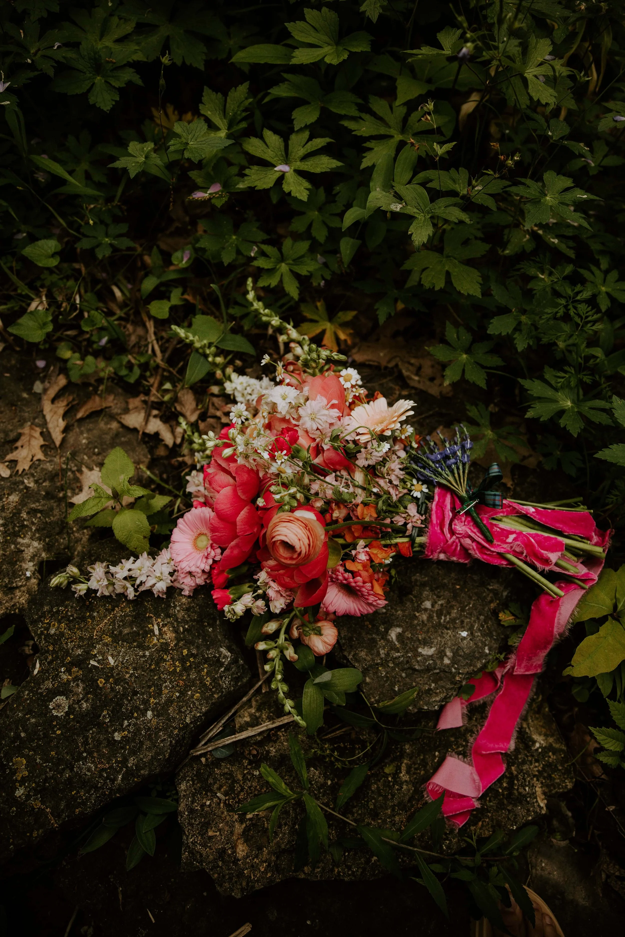 The bridal bouquet full of a variety of pink flowers tied with a hot pink ribbon lays on a rock wall.