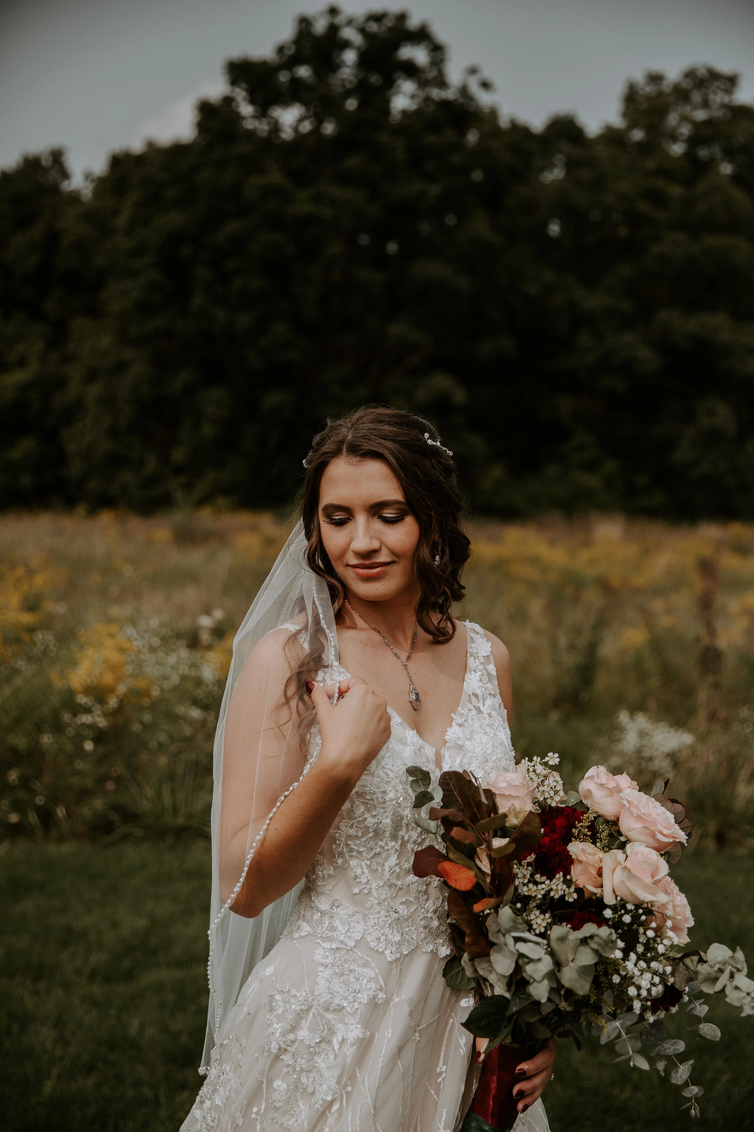 Bride standing in a field, gently holding her veil and bouquet, with a serene expression as she looks down.