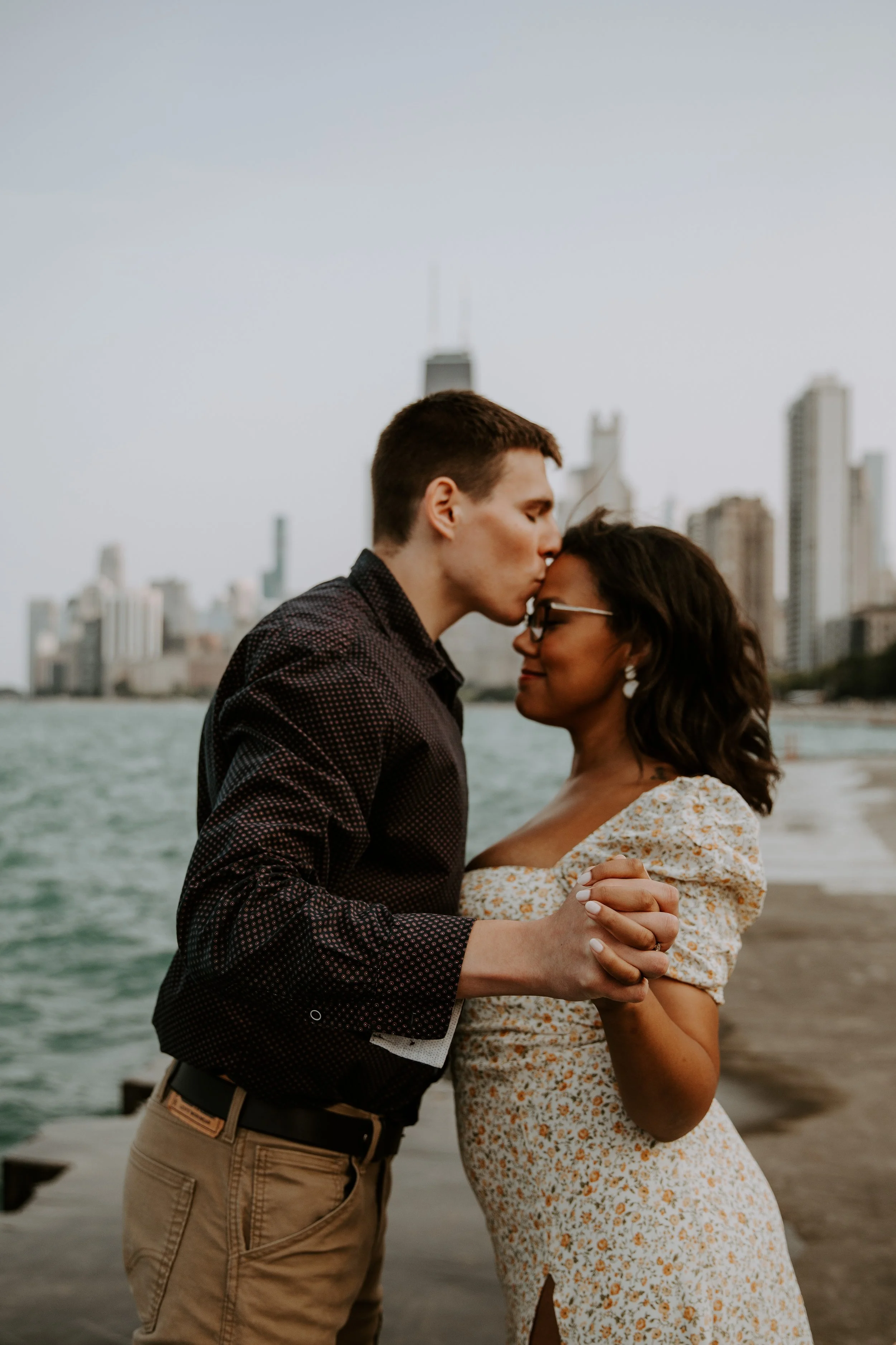 Groom-to-be kissing his fiancée on the forehead as they hold hands by the lake.