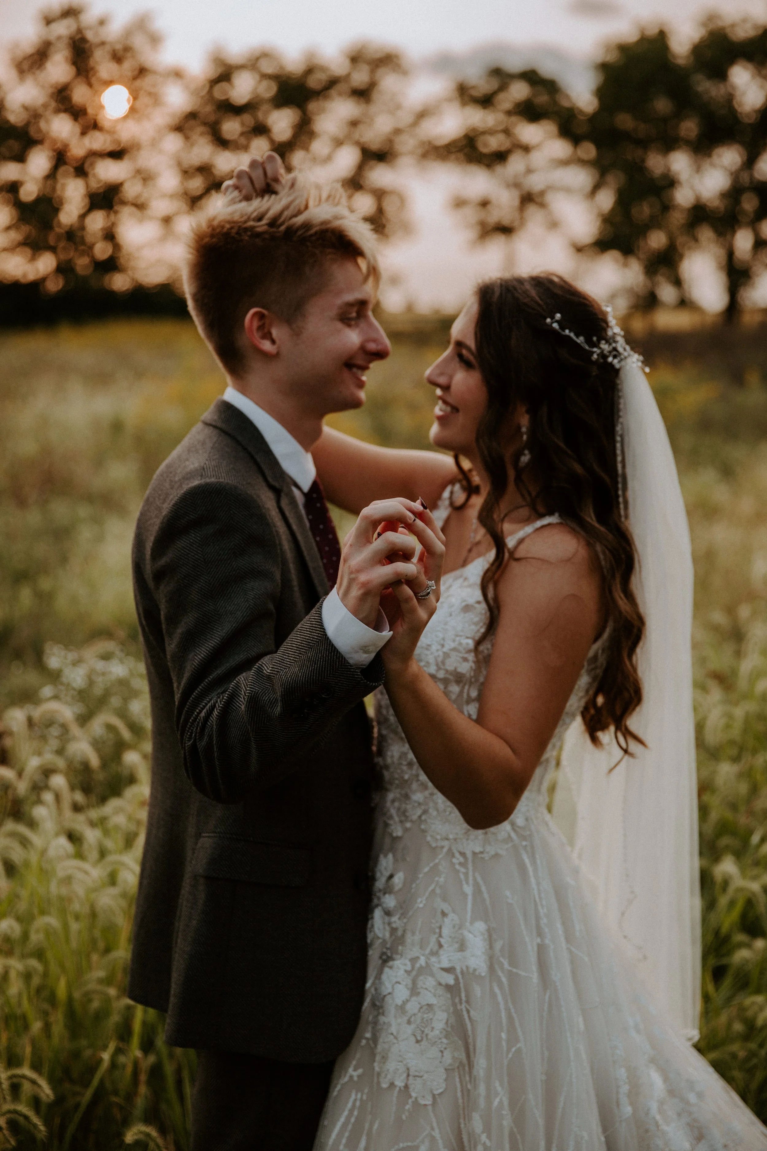 Bride and groom gaze lovingly at each other, smiling as they hold hands in a field during sunset.