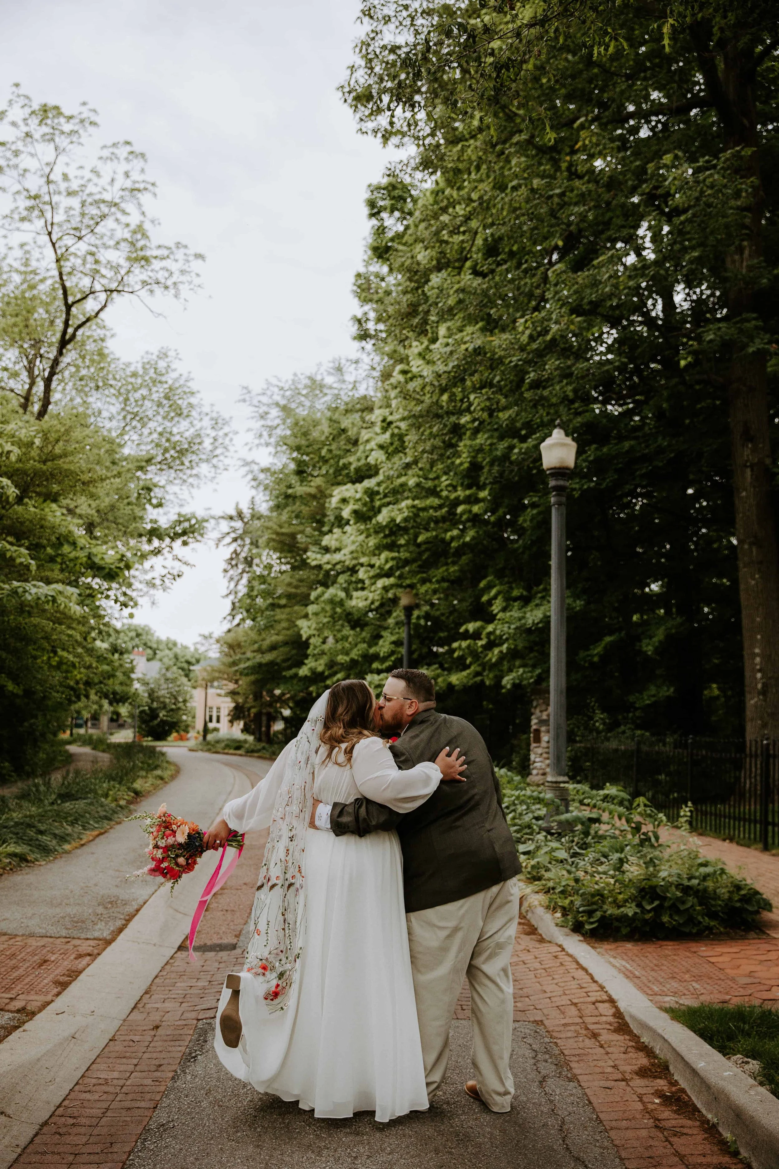 The couple kiss with their backs to the camera. The bride is holding out her flowers and has her leg popped.