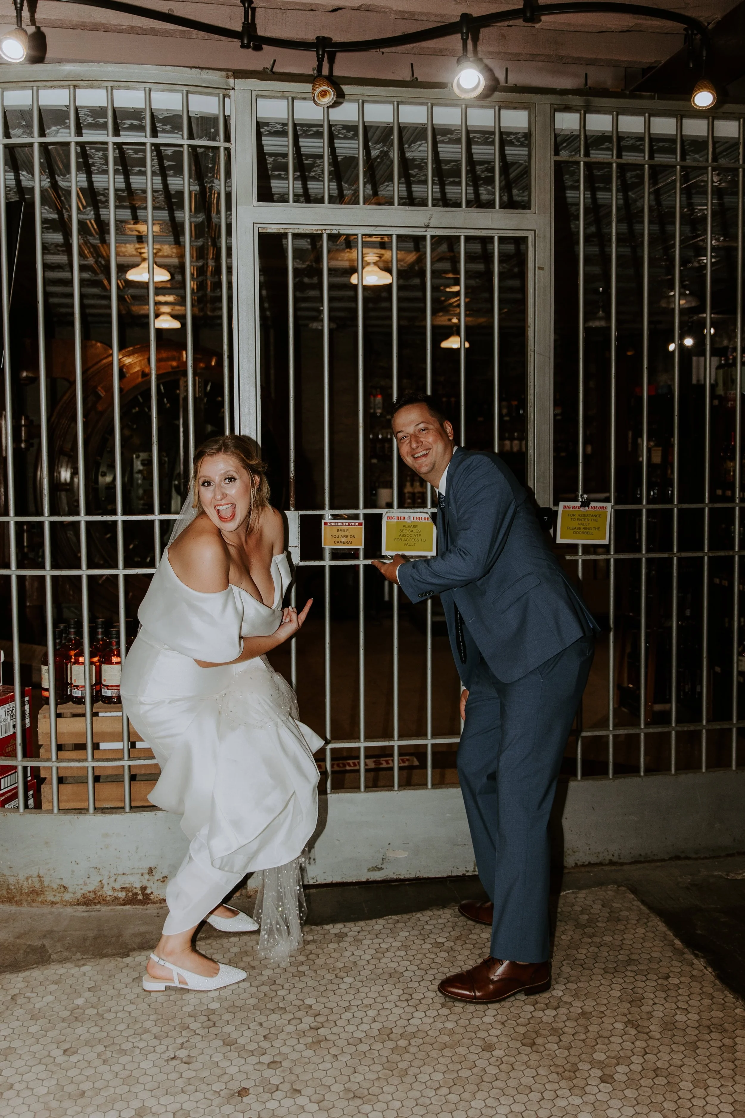 The bride and groom smile big while pointing to a "Smile, you're on camera" sign.