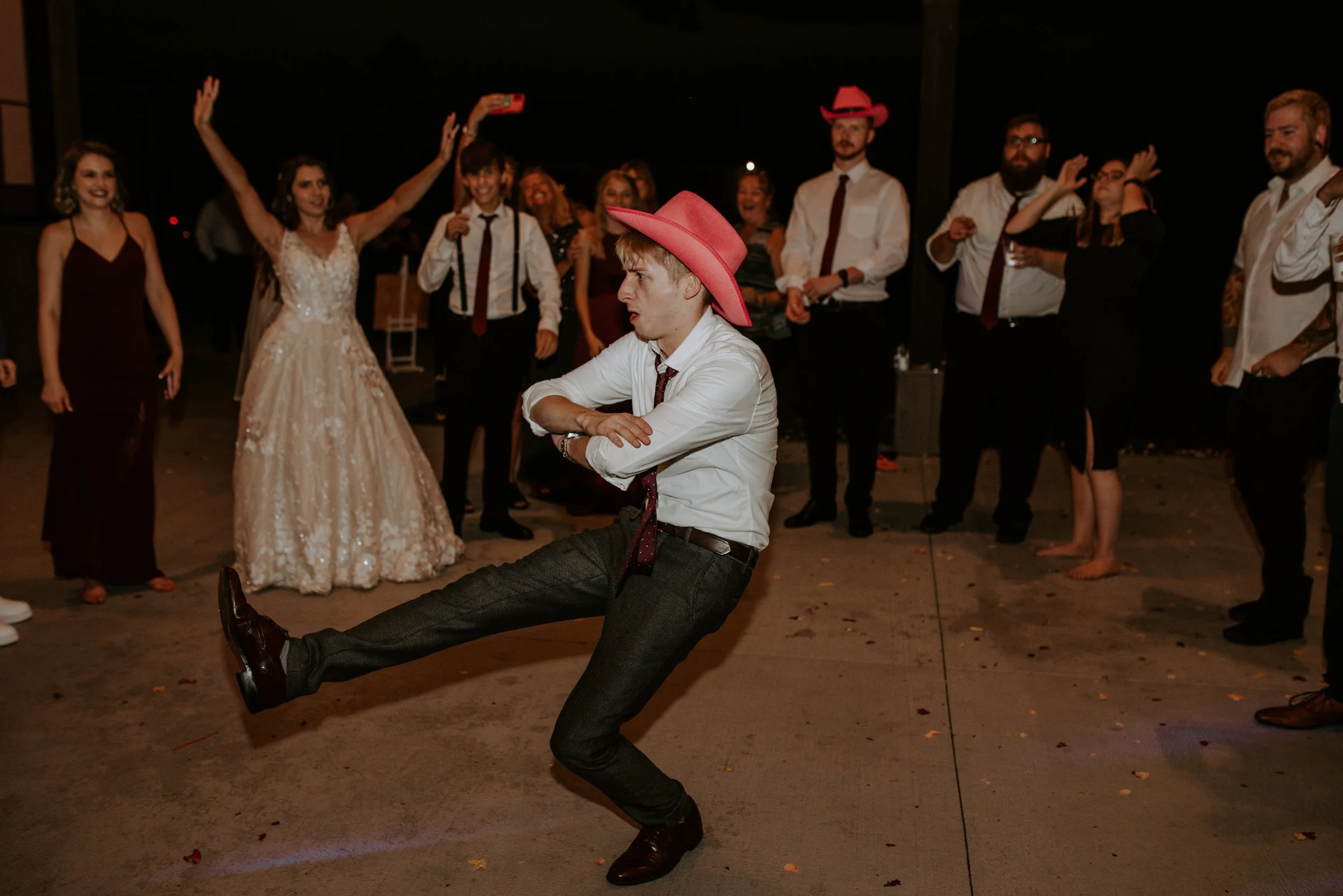 Groom wearing a pink cowboy hat, energetically dancing in the middle of a circle of guests, with the bride cheering him on.