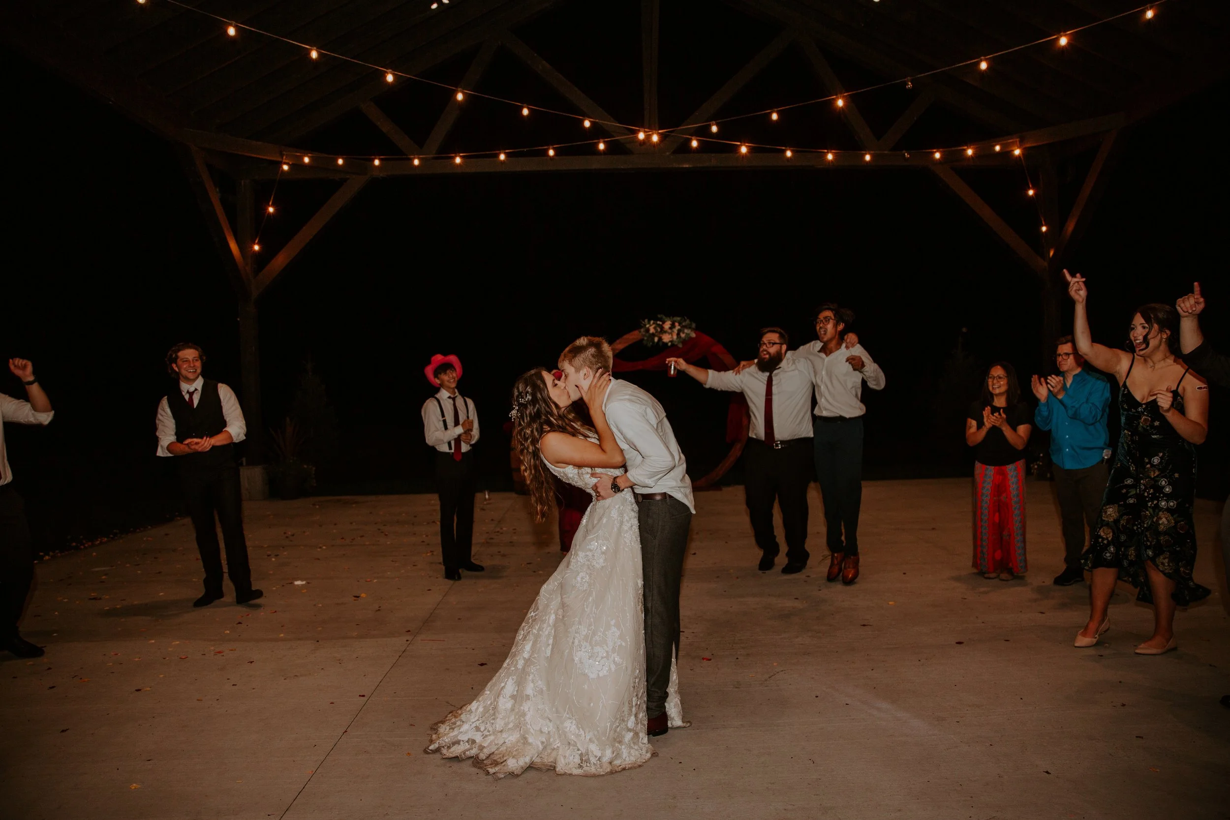 Bride and groom share a romantic kiss on the dance floor under an illuminated pavilion, with guests celebrating around them.