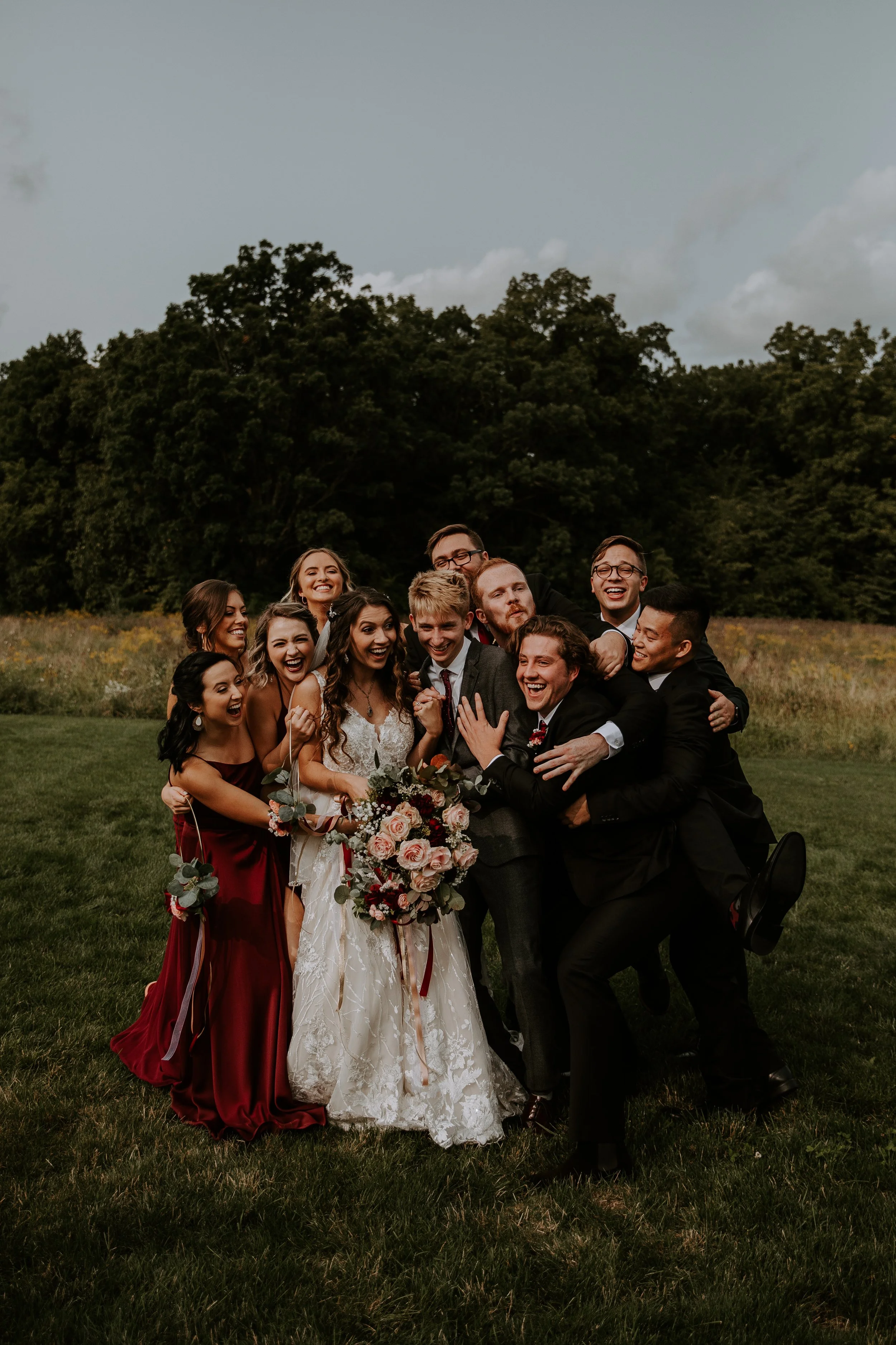 Bride and groom surrounded by their joyful wedding party, all laughing and embracing in a celebratory group hug outdoors.