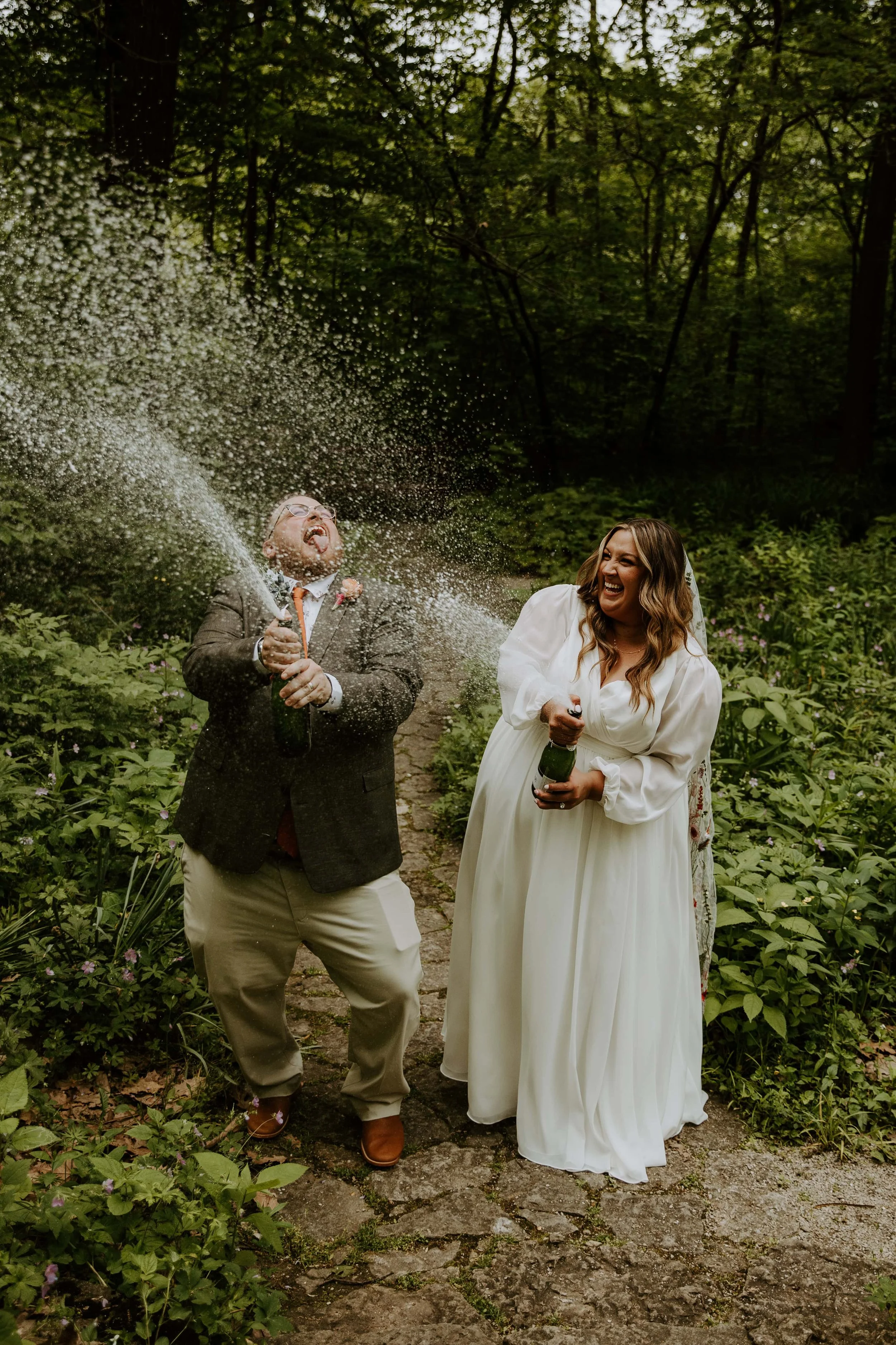 The bride and groom laugh while they each pop a bottle of champagne.