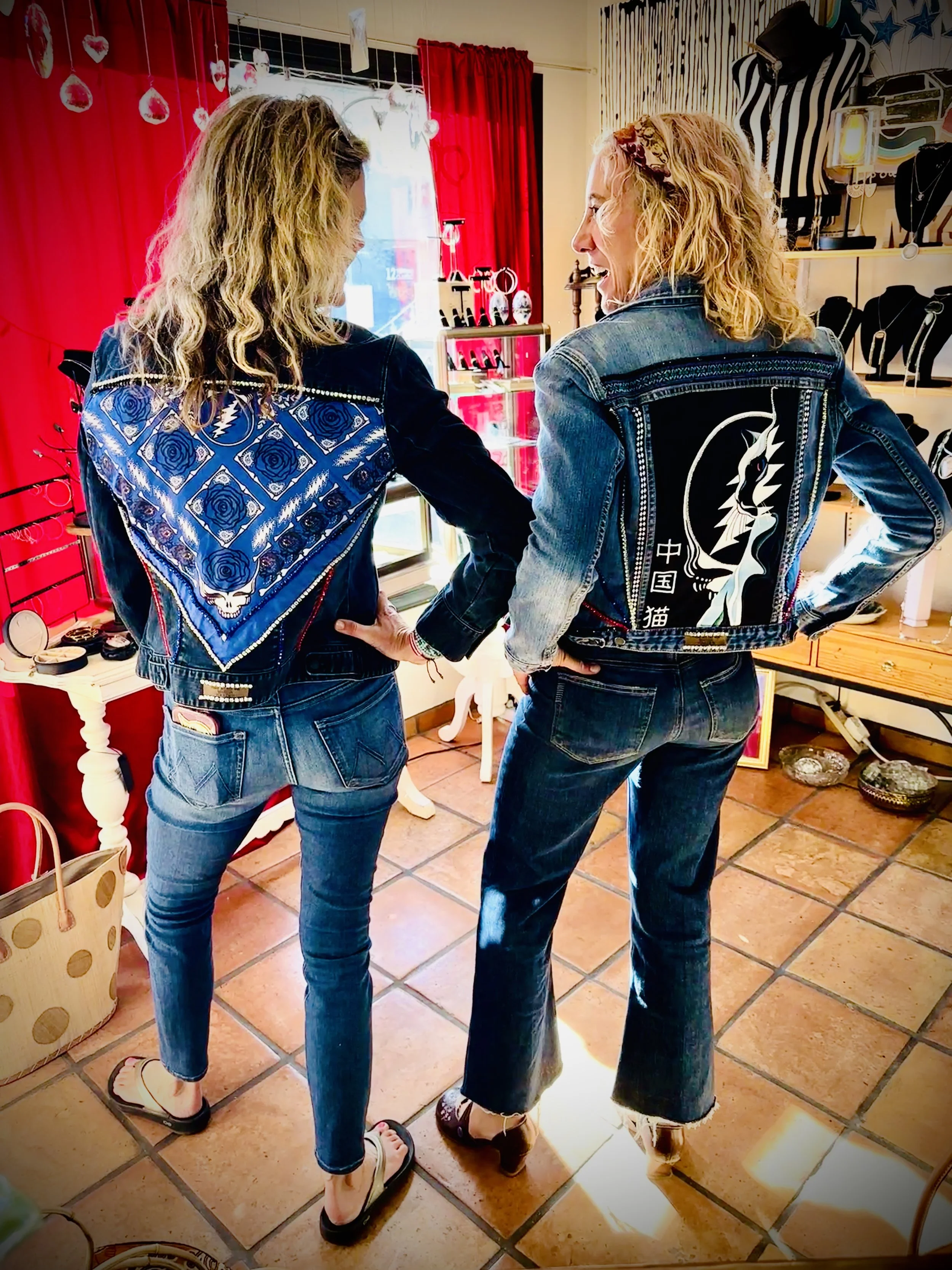 Two women wearing custom denim jackets with designs on the back, standing in a store with red curtains and accessories on display.