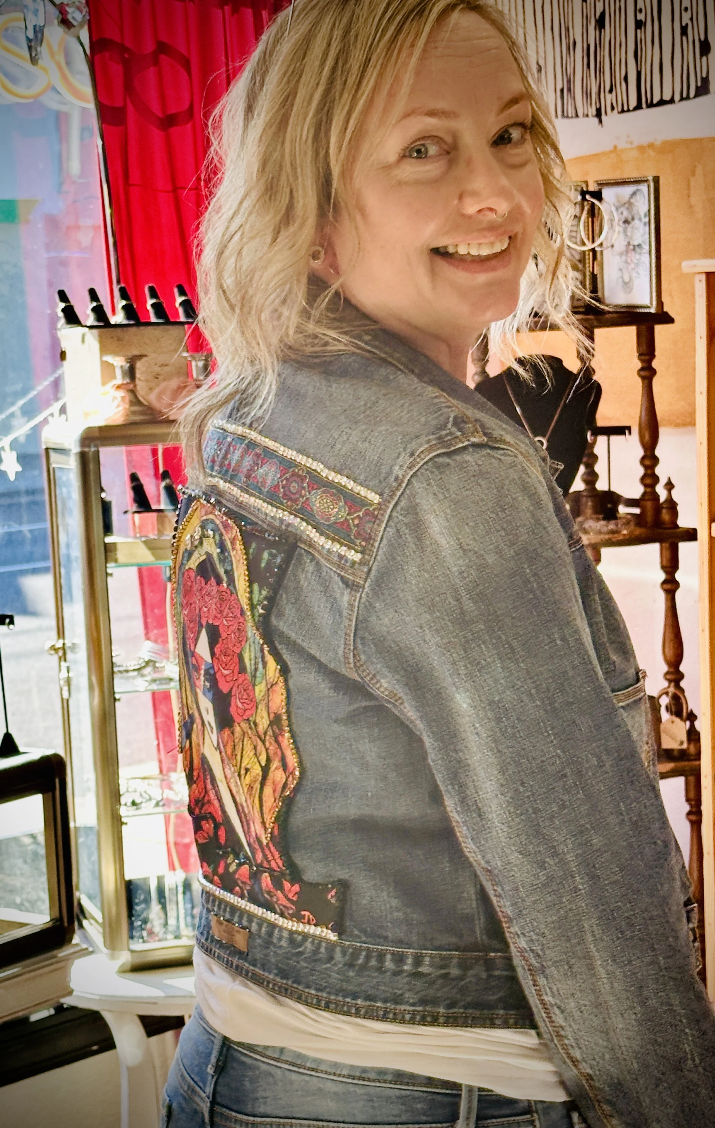 Woman wearing embellished denim jacket with artistic design on the back, standing indoors.