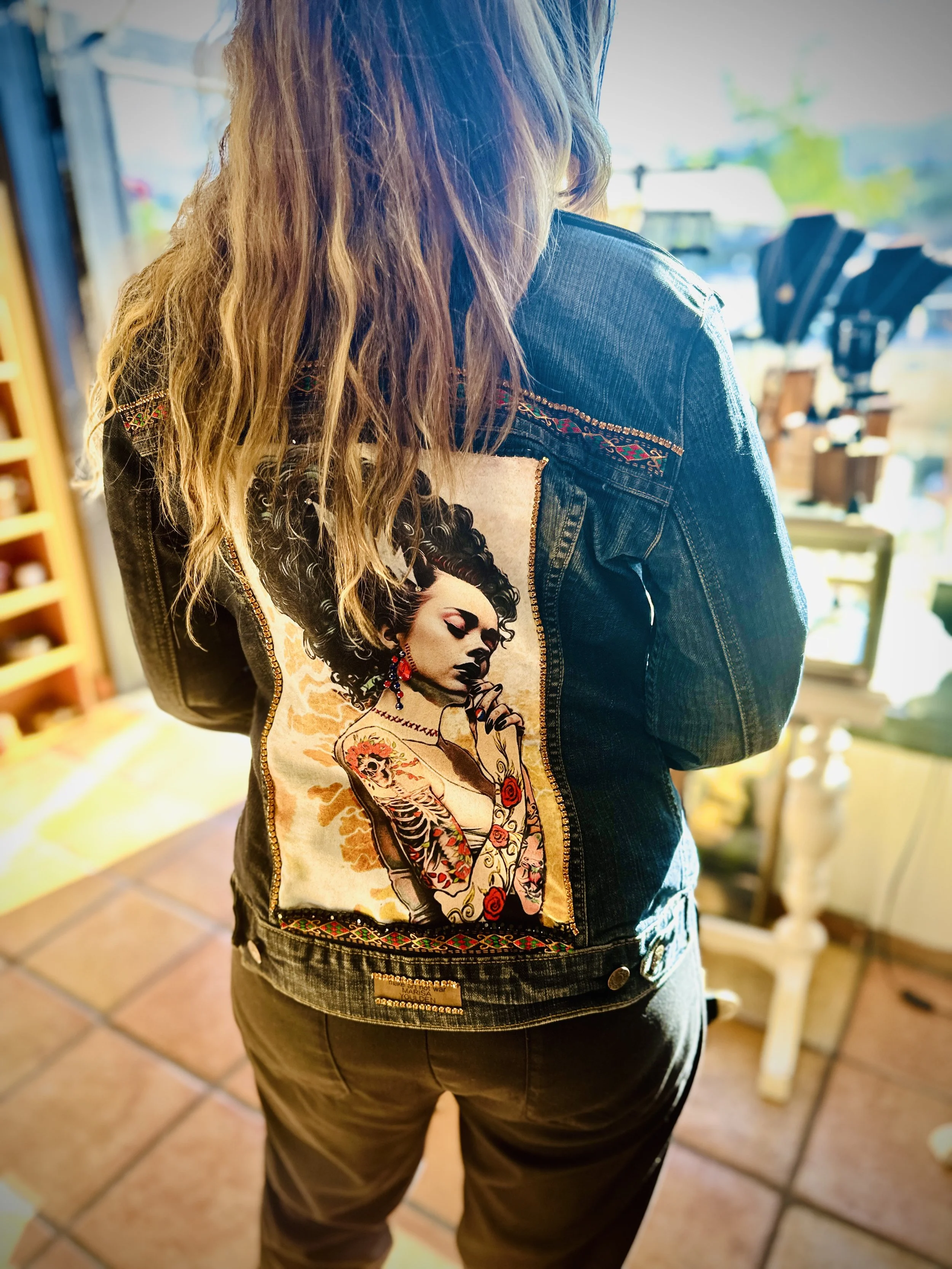 Person wearing a denim jacket with a colorful patch on the back featuring tattoo-style art of a woman with floral tattoos, indoors.