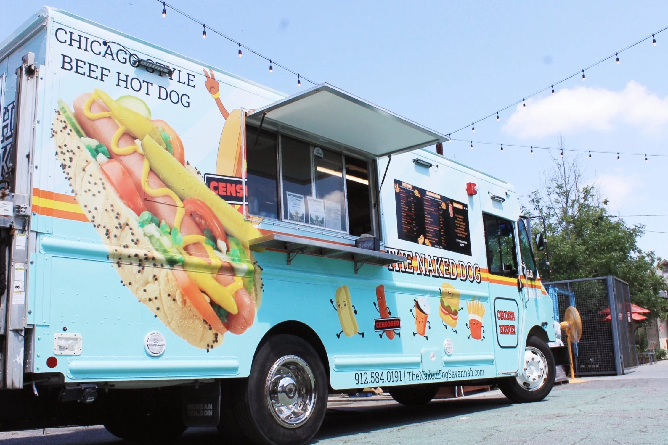 A food truck named 'The Naked Dog' with colorful cartoon hot dog and other fast food drawings on the side, serving hot dogs and quick snacks in an outdoor setting with string lights overhead.