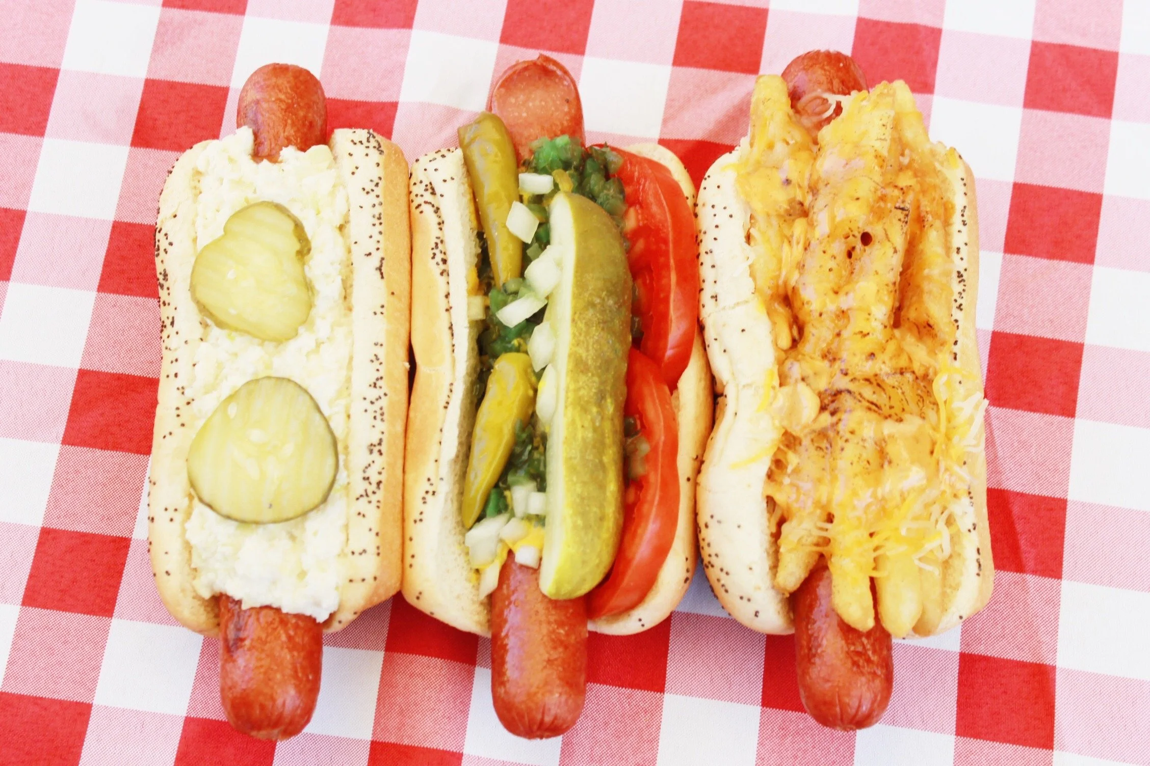 Three hot dogs served in buns with various toppings, placed on a red and white checkered tablecloth. The hot dogs have sausages inside, with toppings including pickles, mustard, onions, tomato slices, shredded cheese, and chili.