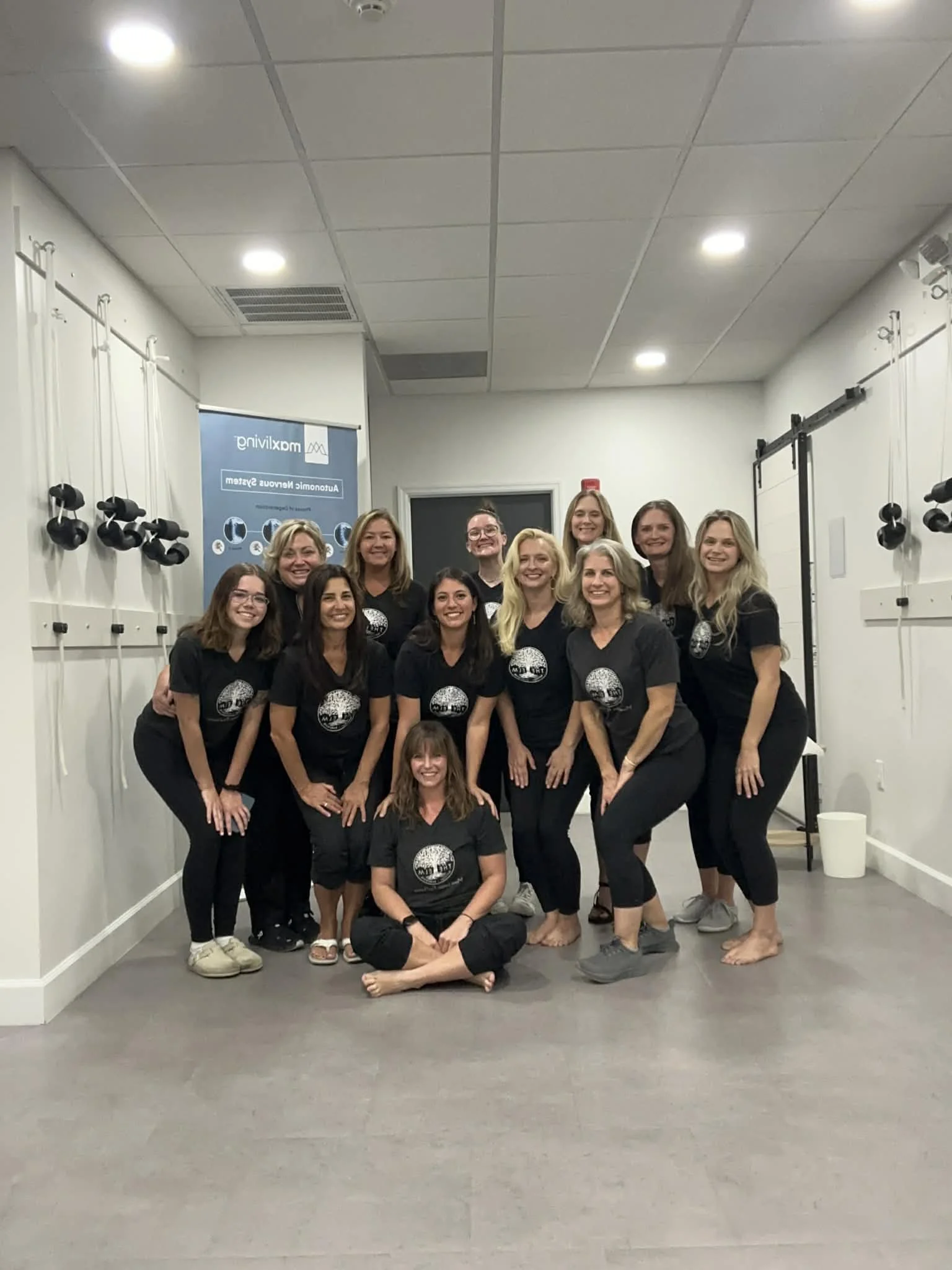 A group of twelve women and one girl, all wearing matching black t-shirts with a circular logo, posing together in a room with white walls and gray flooring. They are smiling and some are crouching or sitting on the floor while others stand behind them.