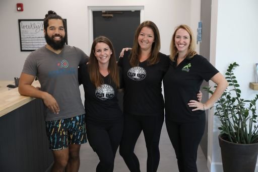 Four people standing together inside a room, smiling at the camera. The group includes one man and three women. They are dressed casually, and the women are wearing matching black shirts with a logo on them. The man is wearing a gray t-shirt and patterned shorts. The room has a potted plant, a whiteboard, and some signs on the wall.