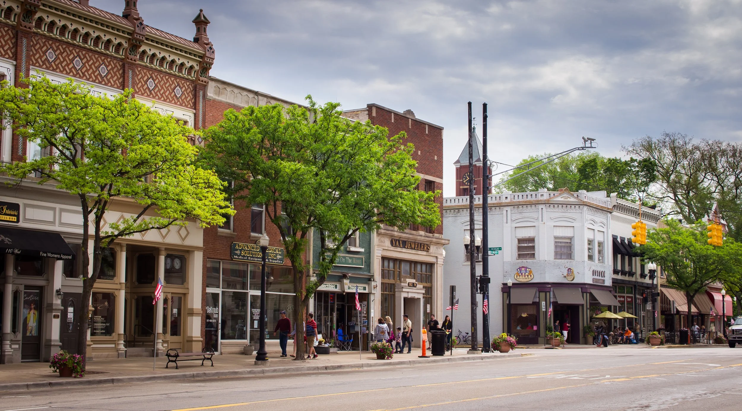 A lively downtown street with storefronts, green trees, and people walking. Buildings feature decorative architecture, and American flags are displayed. Overcast sky overhead.