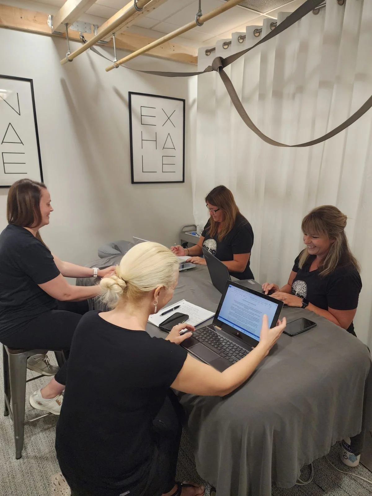 Four women sitting at a table, some using laptops and one taking notes, inside a room with white walls, framed artworks that spell out 'EXHALE' and 'INHALE', and massage equipment hanging from the ceiling.
