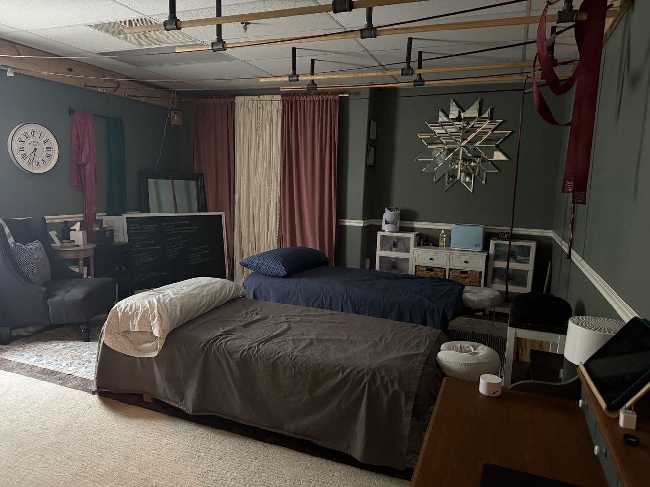 A room with two massage tables, and various furniture including a black armchair, a white cabinet, and a desk with a tablet, speaker, and other items. Colors include green walls, multicolored curtains, and ceiling-mounted wooden and metal fixtures.
