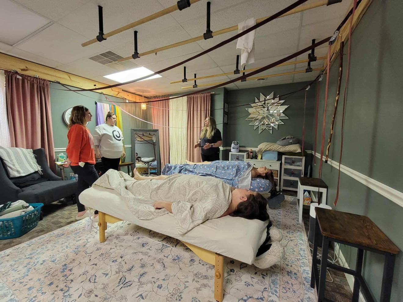A room with two women lying on massage tables and two women standing and talking, with safety straps and ashiatsu bars suspended from the ceiling, and colorful curtains and decor.