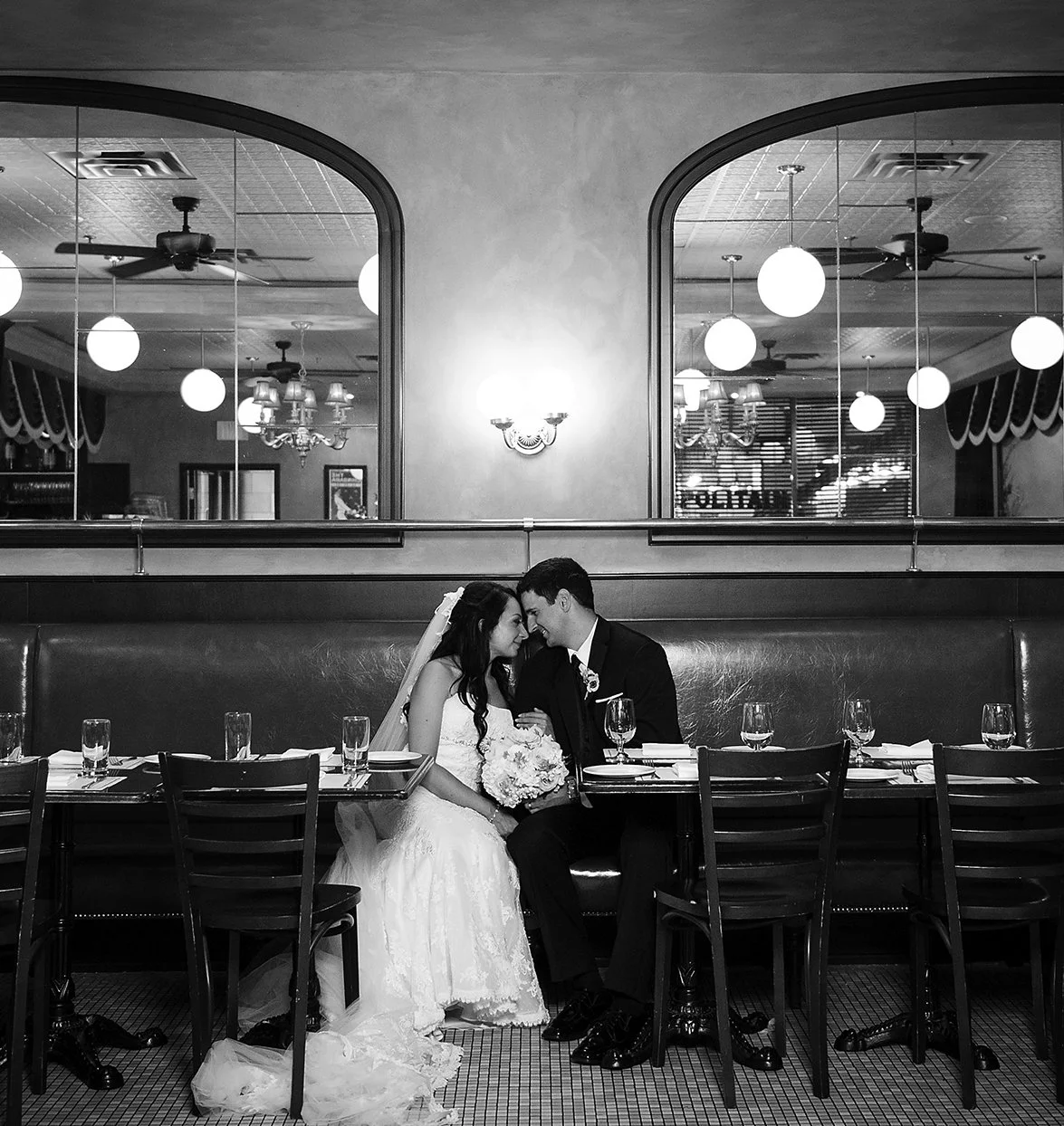 Black and white photograph of a bride and groom sitting together at a restaurant table, touching foreheads and smiling, with empty wine glasses and plates in front of them, and large windows with hanging globe lights and fans in the background.