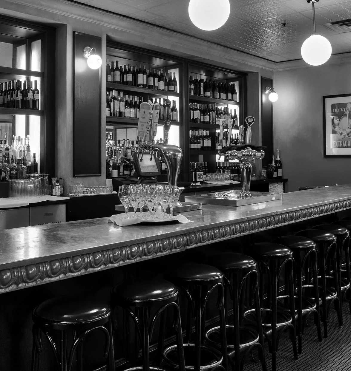 Black and white photo of a bar with bottles of wine on shelves, glasses on the counter, and bar stools in front.