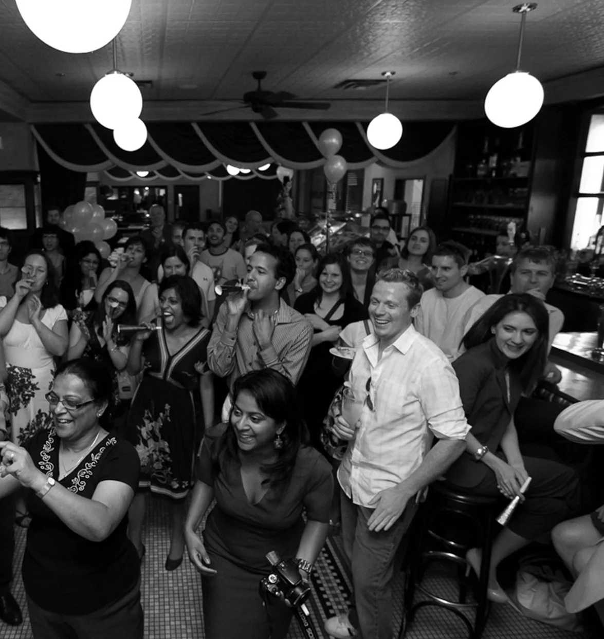 Group of people celebrating at a party, some holding drinks and taking photos, in a decorated indoor venue with balloons and hanging lights.