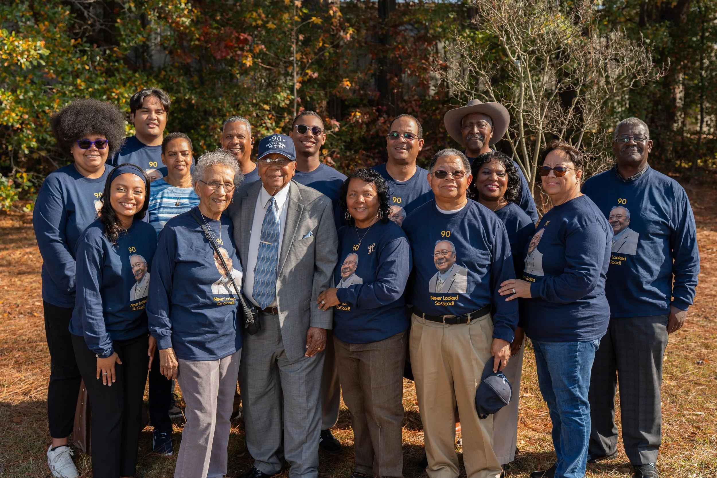 Group of diverse people standing outdoors on a sunny day, wearing navy blue shirts with a printed image and text, some also have sunglasses and hats, with autumn trees and fallen leaves in the background.