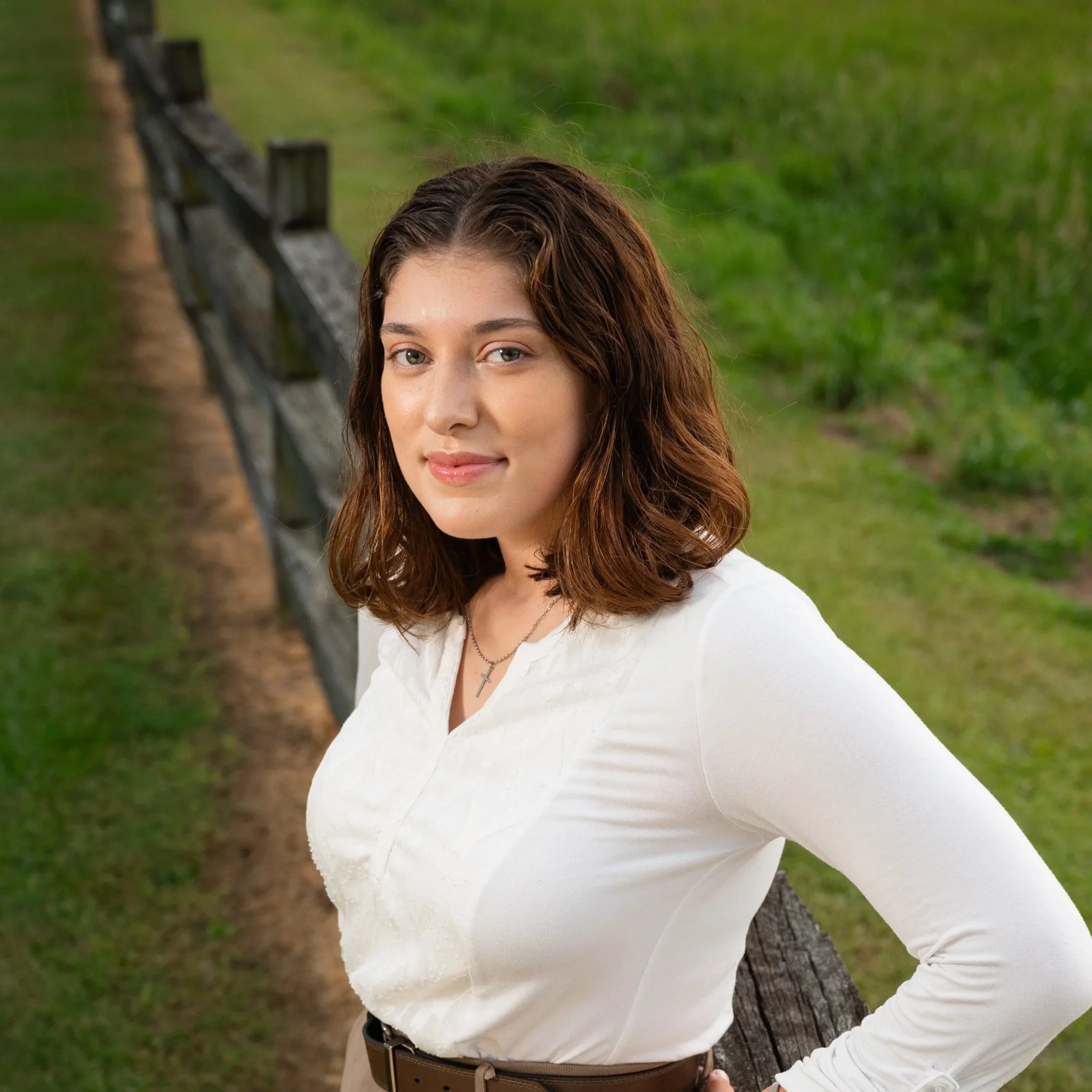 A young woman with shoulder-length brown hair and fair skin, wearing a white blouse and a cross necklace, standing outdoors next to a wooden fence with green grass and plants in the background.