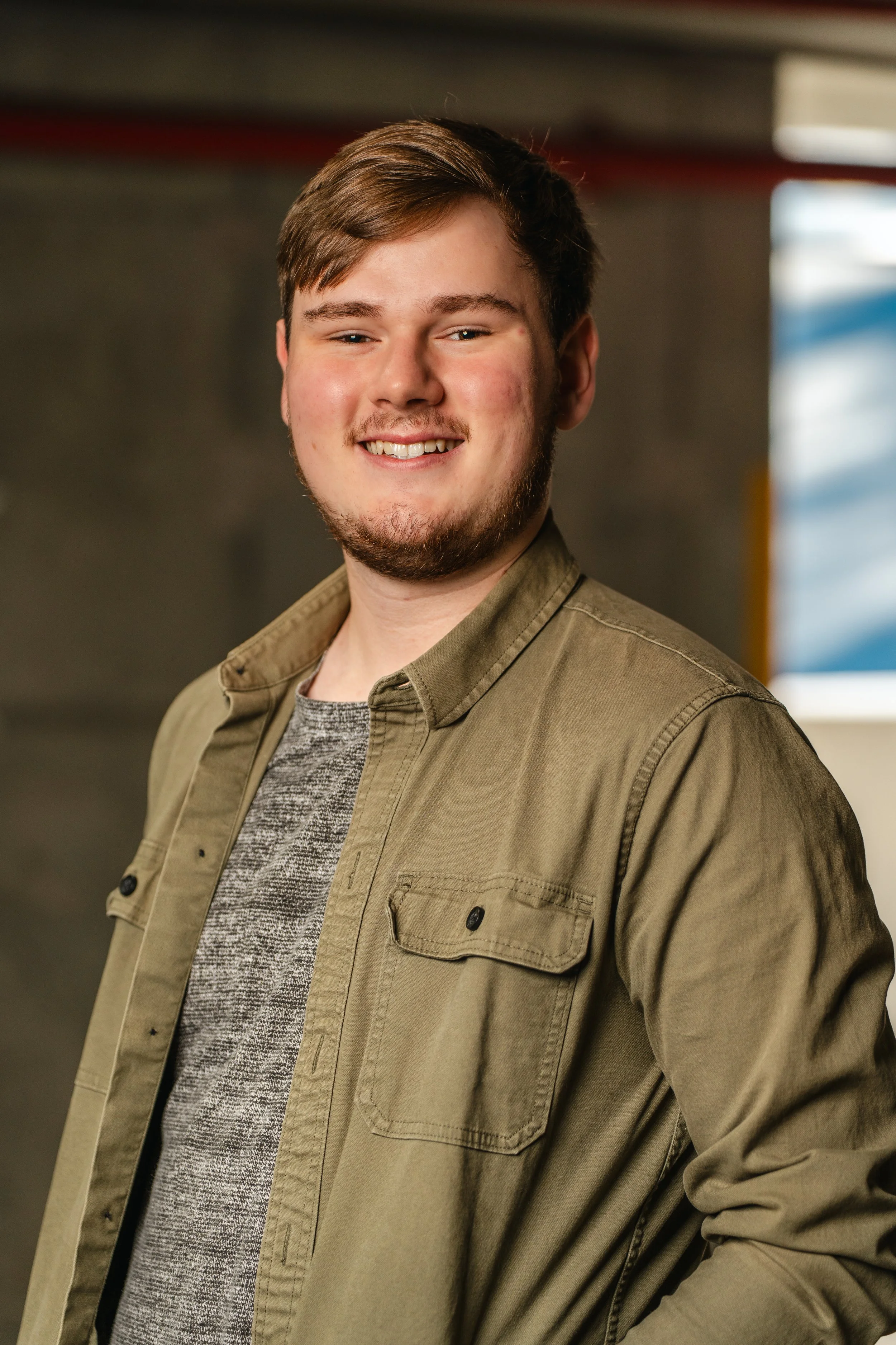 A young man with short brown hair, beard, and mustache, smiling, wearing a green shirt over a gray sweater, standing against a blurred background.