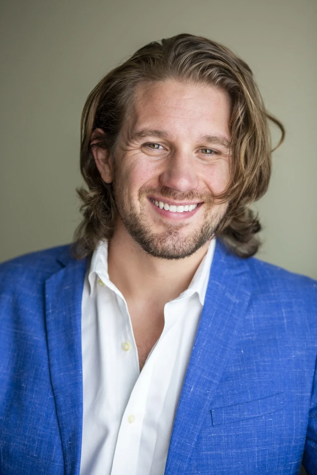 Headshot of Michael Alder smiling with long, wavy brown hair, wearing a blue blazer and a white shirt, against a neutral background.