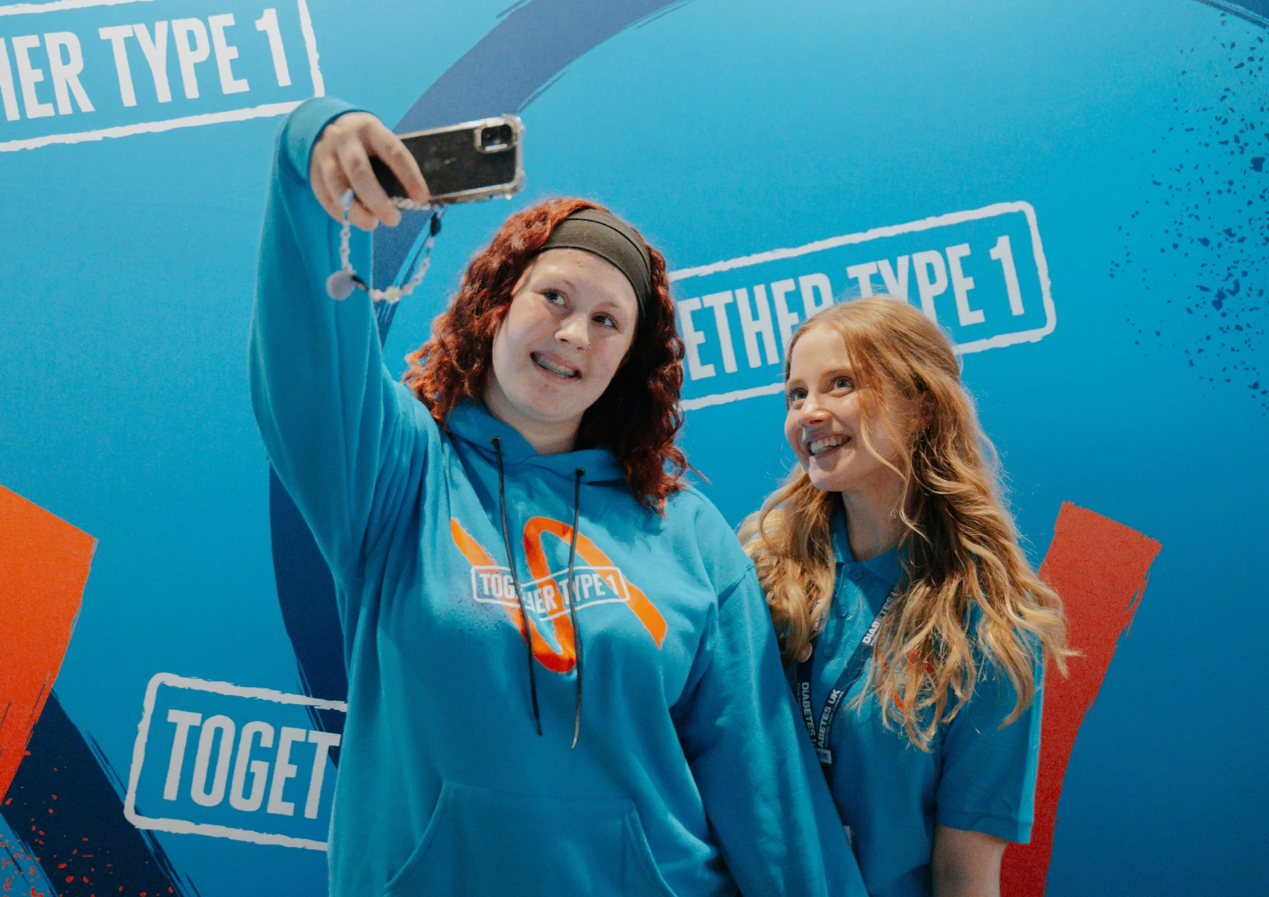 Women taking selfie in front of Together Type 1 backdrop wearing branded hoodies