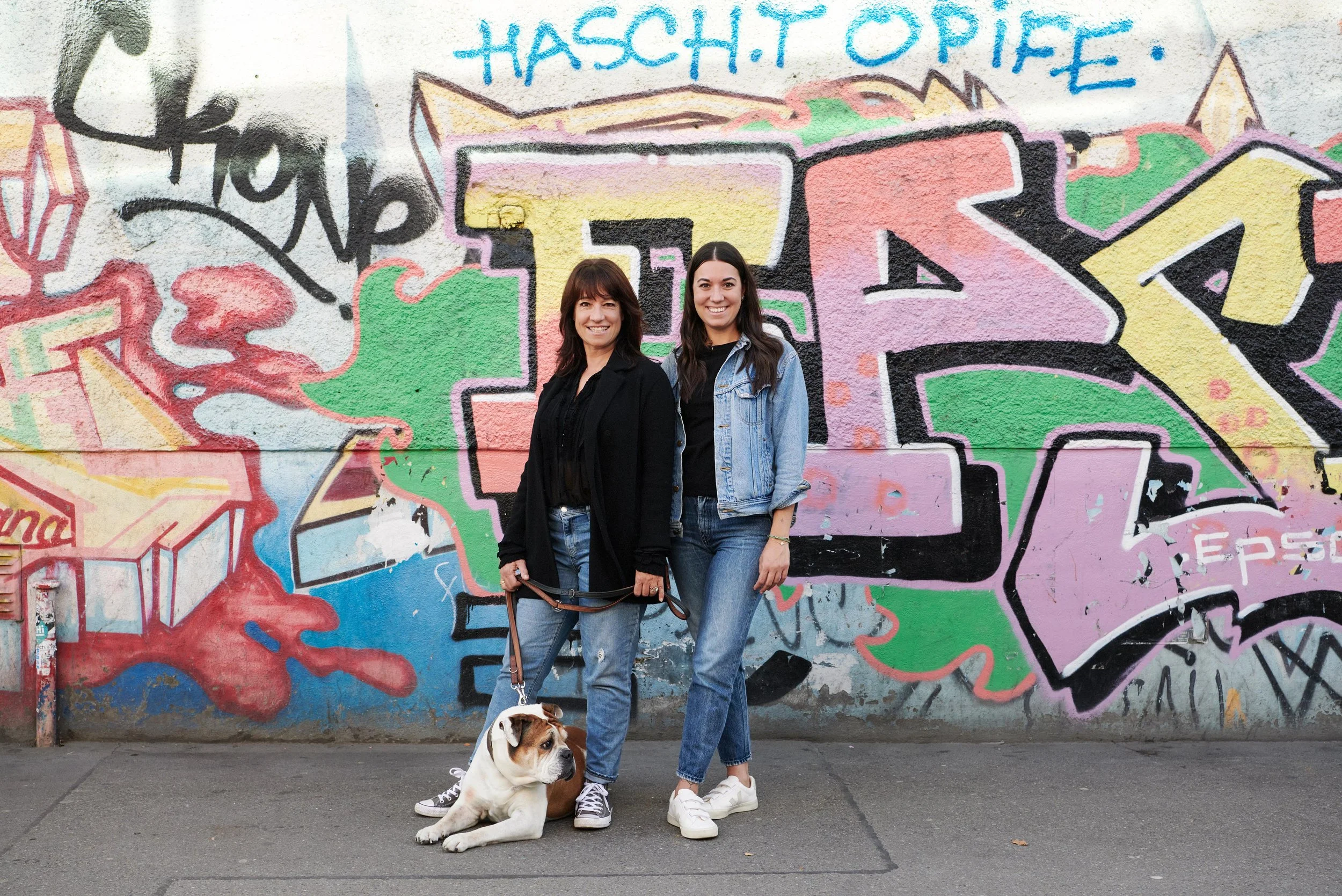 Zwei Frauen und ein Hund stehen vor einer bunten Graffiti-Wand. Eine Frau hält die Leine eines sitzenden Hundes. Das Graffiti enthält große, farbige Buchstaben und Wörter, darunter 'HASS', 'OPF' und 'E'.