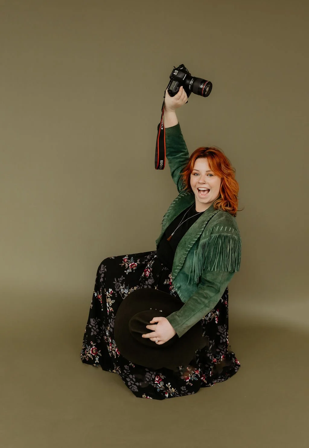 A women with red wavy hair crouched down holding camera over her head in fringe jacket
