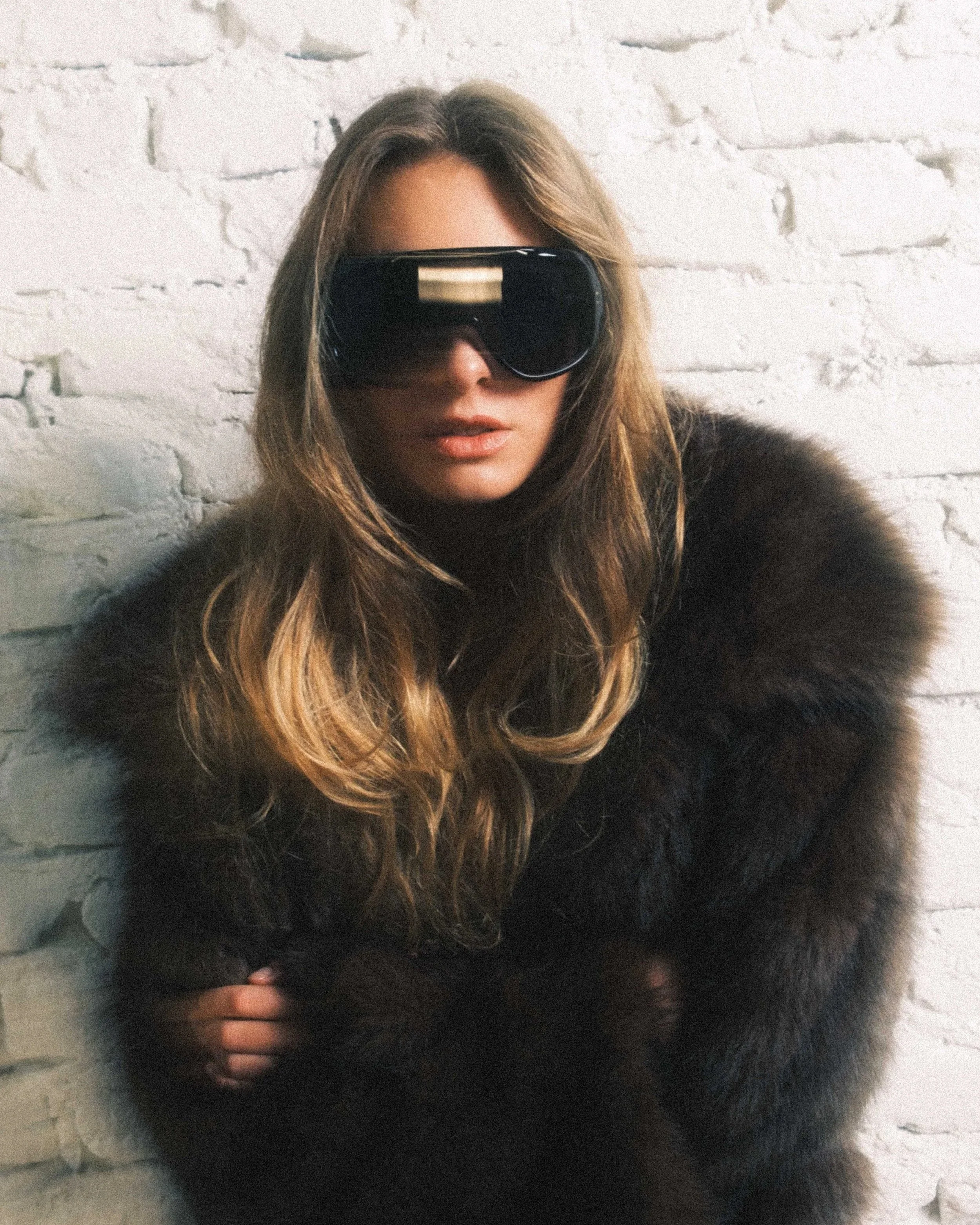 Frau mit großen schwarzen Sonnenbrillen und Felljacke vor einer weißen Ziegelwand.
