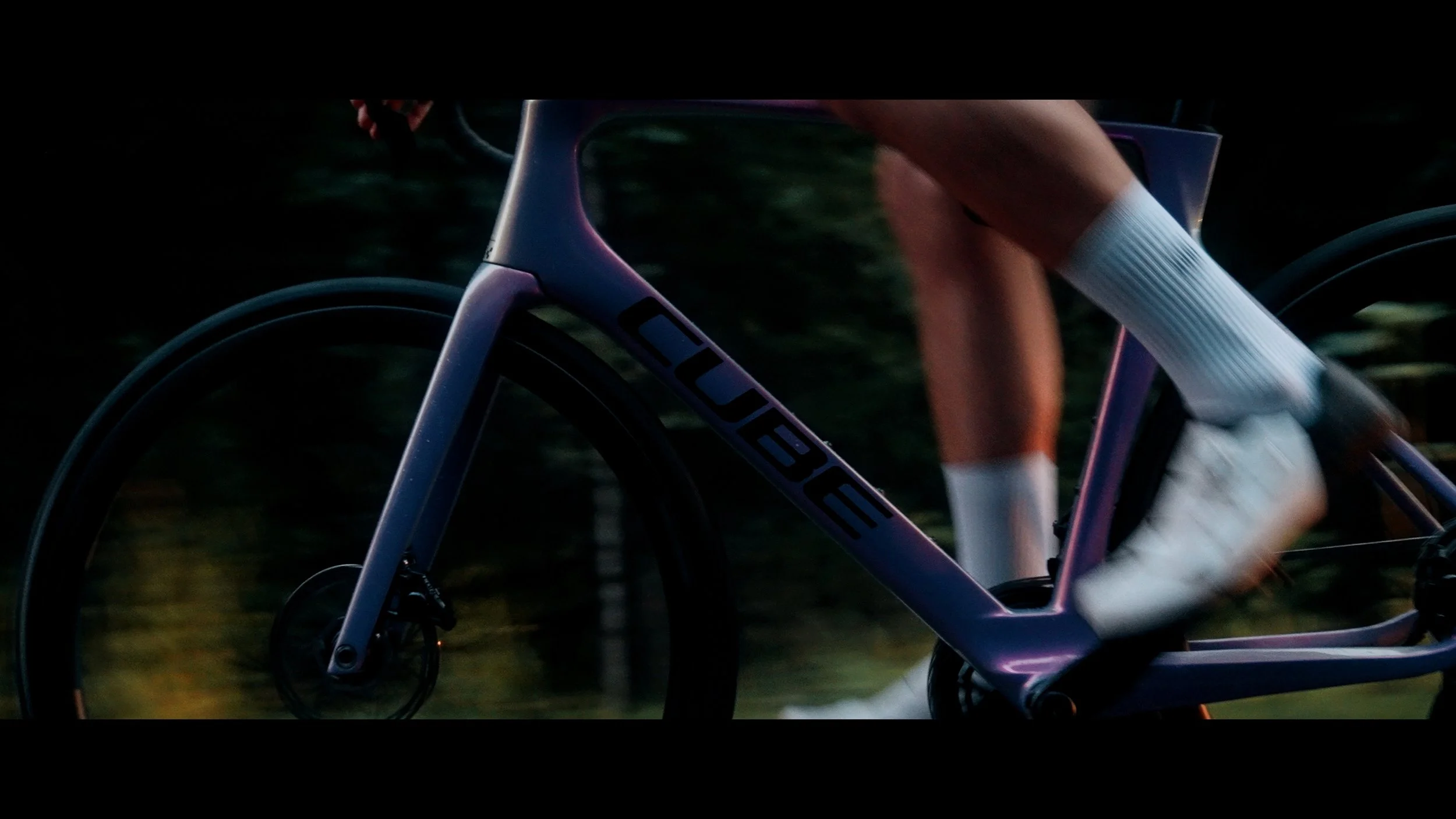 Nahaufnahme eines stilvollen, modernen Rennrads mit dem Markennamen 'CUBE' auf dem Rahmen, während eine Person in weißen Sportsocken und Laufschuhen auf einem Camping- oder Waldweg fährt, umgeben von Natur bei Sonnenuntergang.