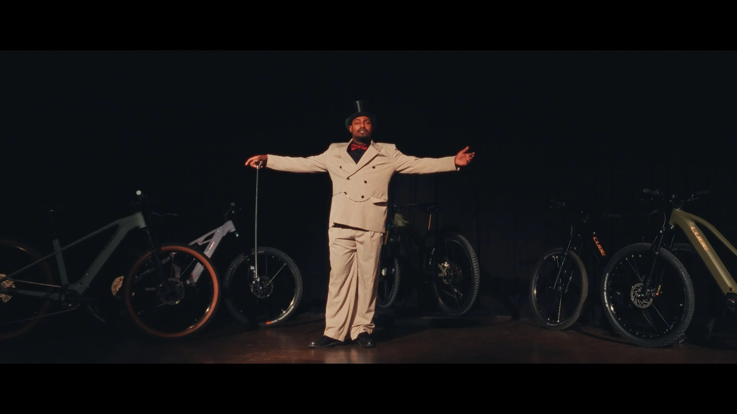Man in Zylinderhut, beige Anzug, roter Fliege, mit ausgestreckten Armen auf einer Bühne, umgeben von Mountainbikes im Dunkeln.