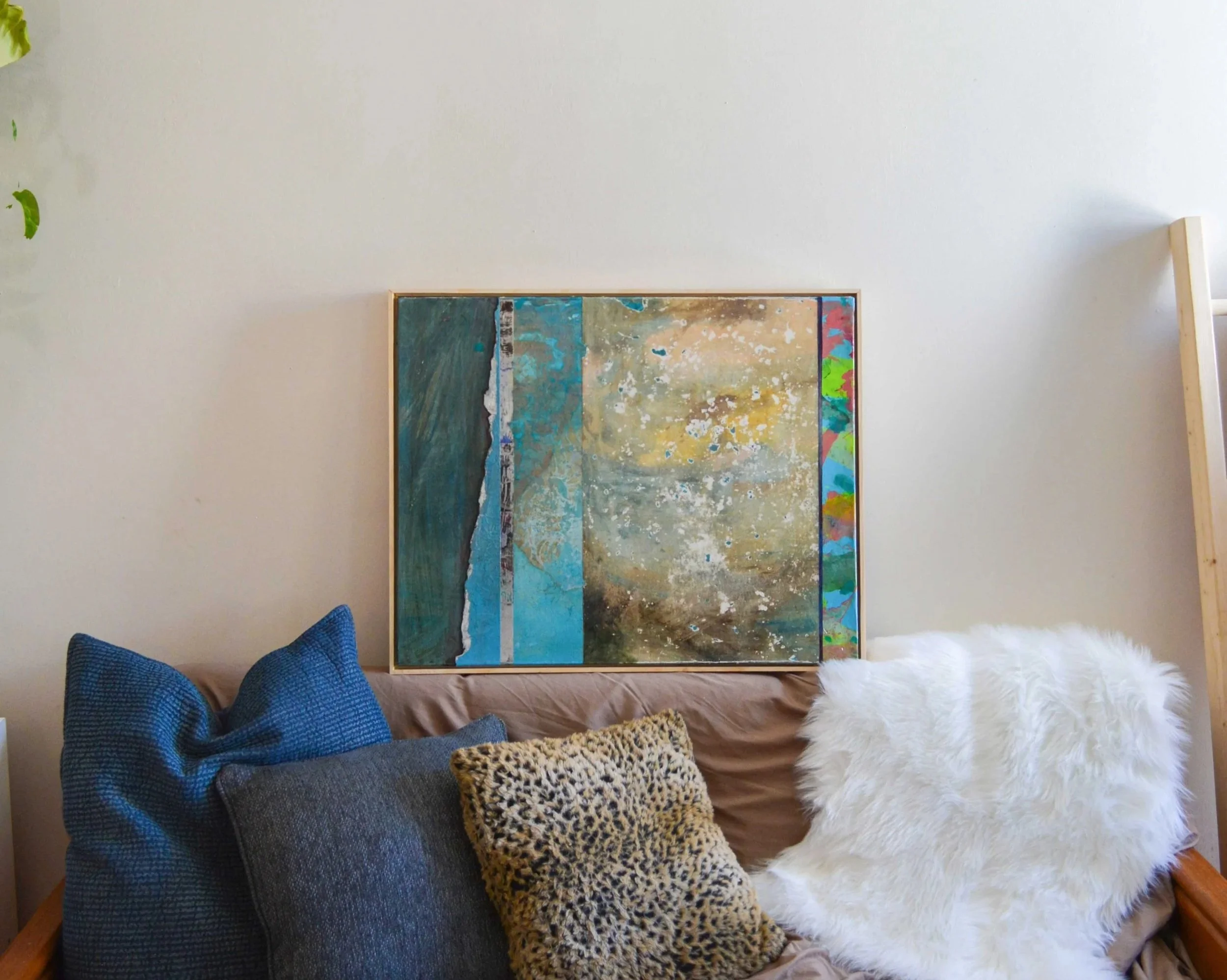 Living room with a beige sofa decorated with blue, gray, leopard print, and white faux fur pillows. A colorful abstract painting is hung on the wall above the sofa. Part of a wooden ladder shelf is visible on the right, and a small green plant is on 
