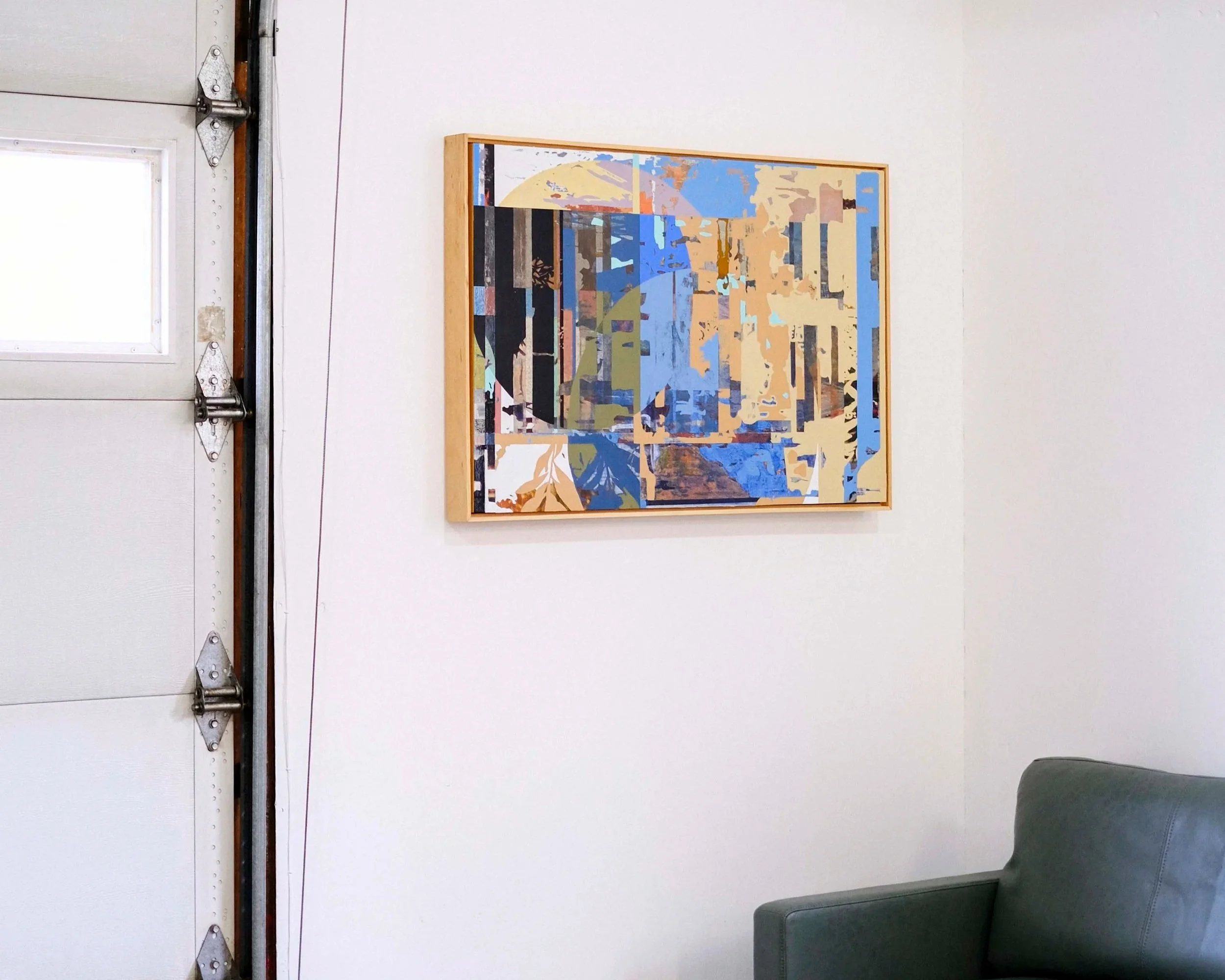 Abstract landscape painting in a wooden frame hanging on a white wall next to a gray couch.