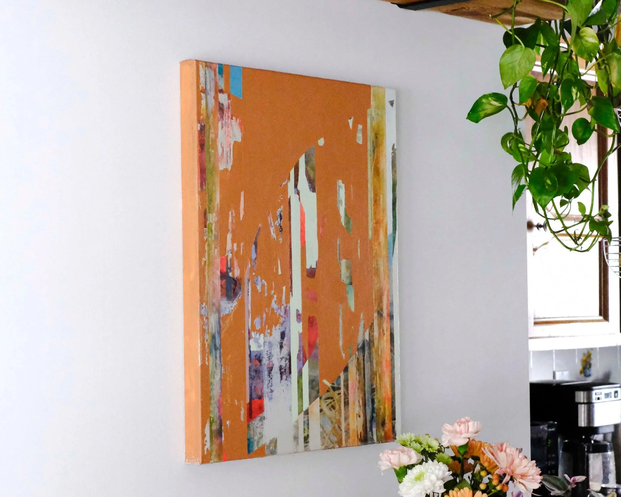 An abstract painting with orange and multi-colored shapes hanging on a light wall in a room with a window and a plant nearby.