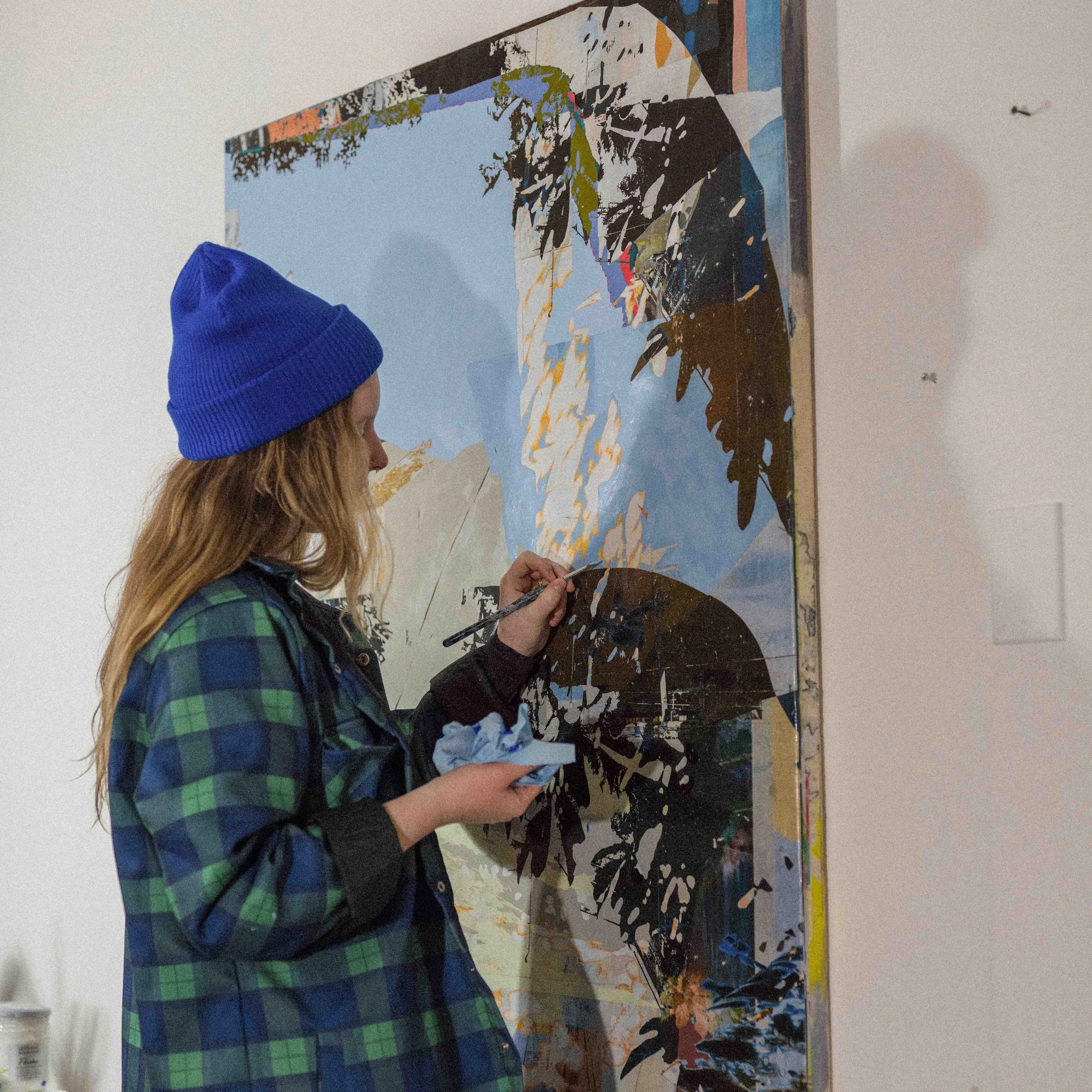 A woman with long hair wearing a blue beanie and a green and blue plaid jacket is painting a large abstract artwork on a wall. She is holding a paintbrush in one hand and a cloth or palette in the other.