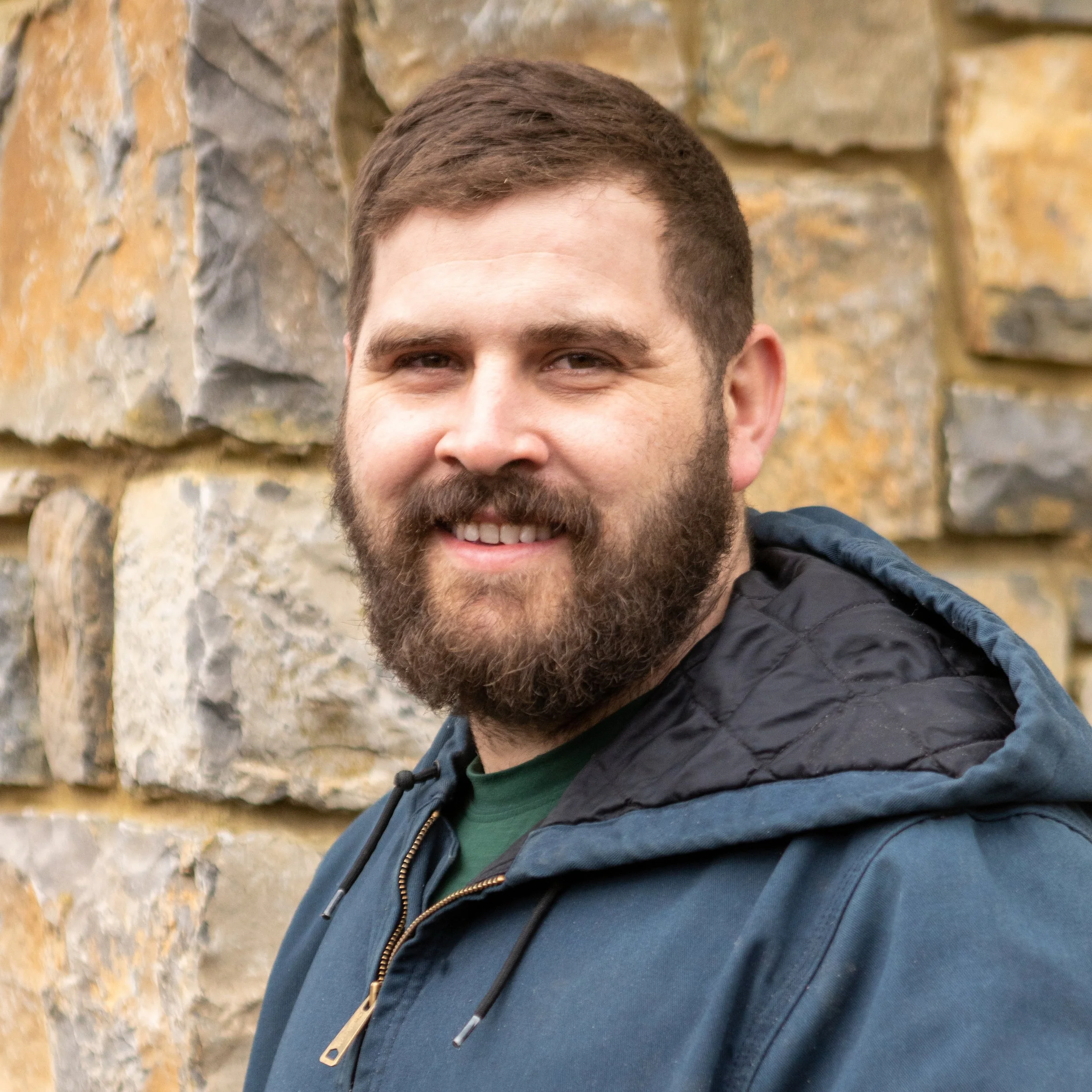 A man with a beard smiling outdoors in front of a stone wall.