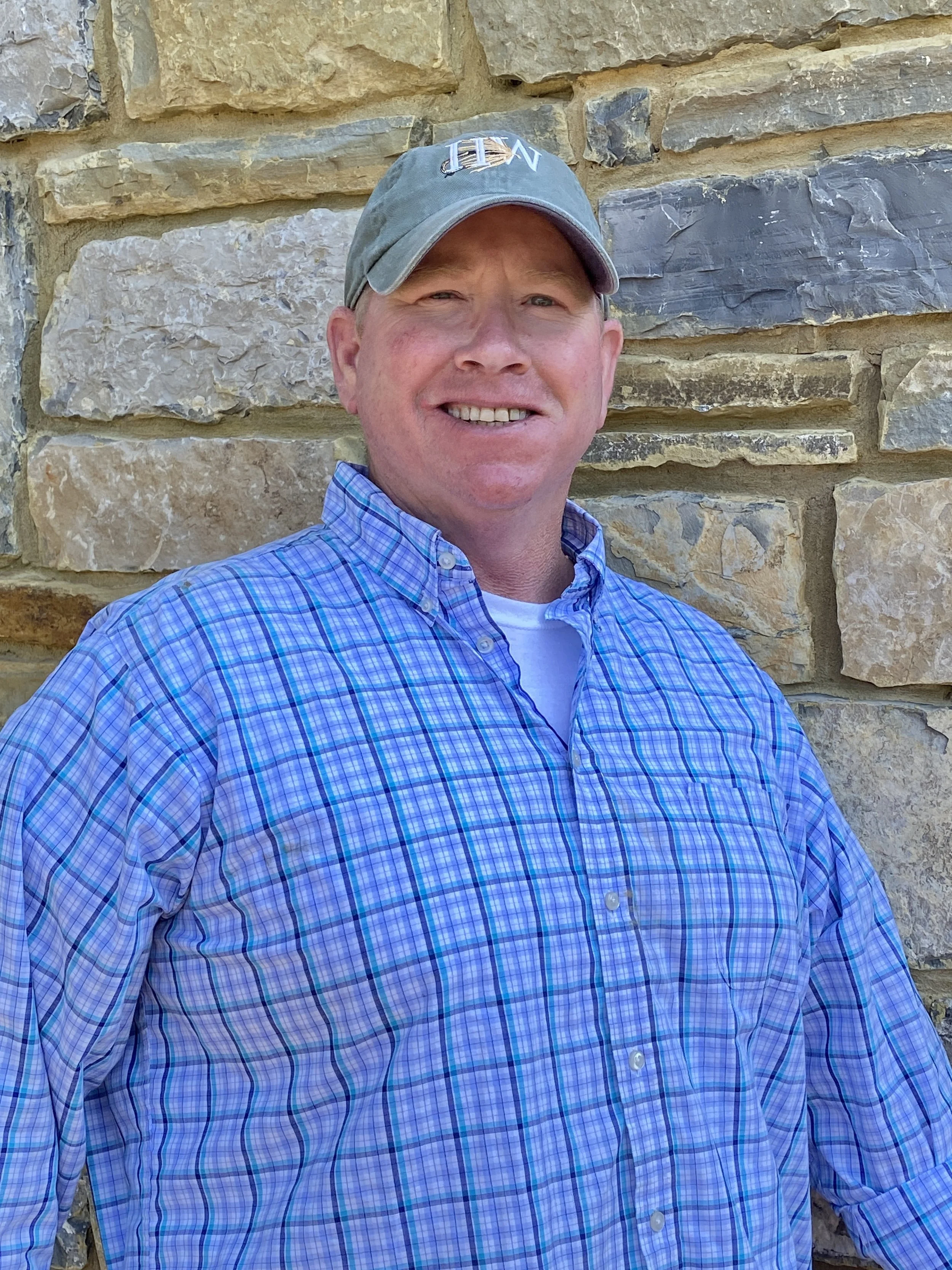 A man outdoors smiling, wearing a gray baseball cap and a blue plaid button-up shirt, standing in front of a stone wall.