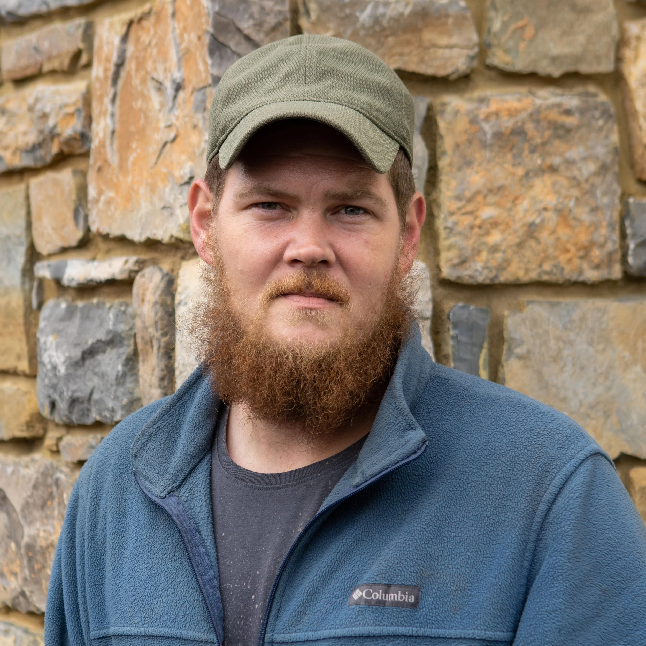 A man with a beard and mustache wearing a green cap and a blue Columbia fleece jacket standing outdoors against a stone wall.