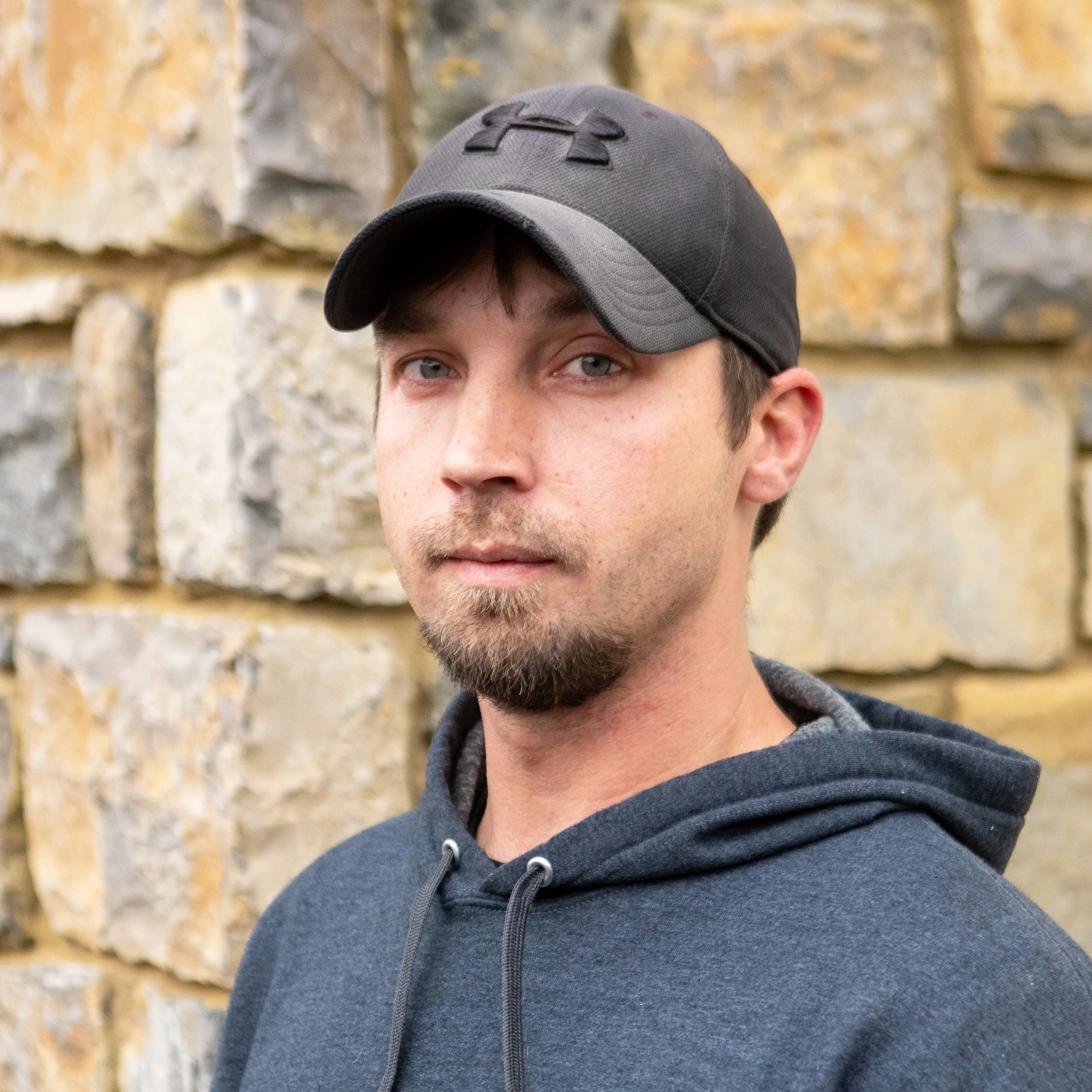 A young man with light skin, wearing a black and gray Under Armour baseball cap and a dark gray hoodie, standing in front of a stone wall.