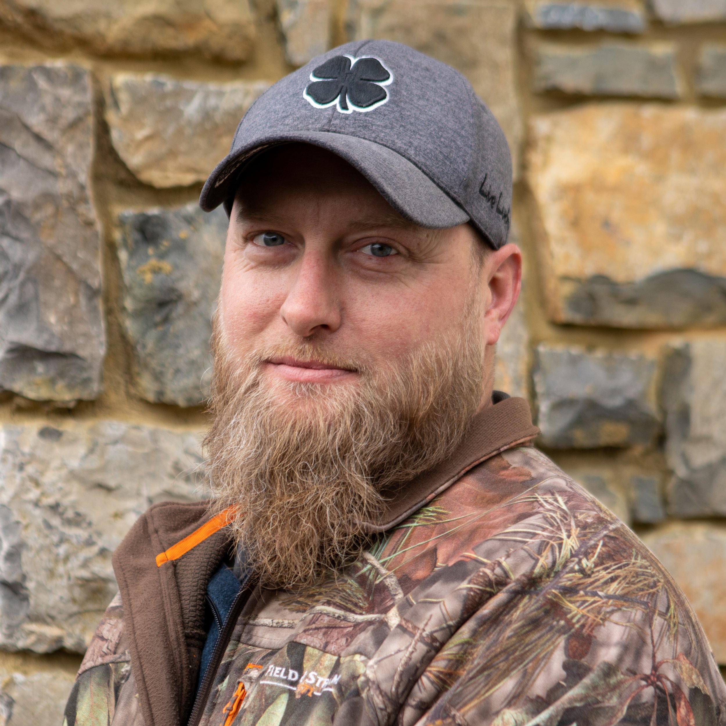 A man with a beard wearing a gray cap with a four-leaf clover patch and a camouflage jacket stands in front of a stone wall.