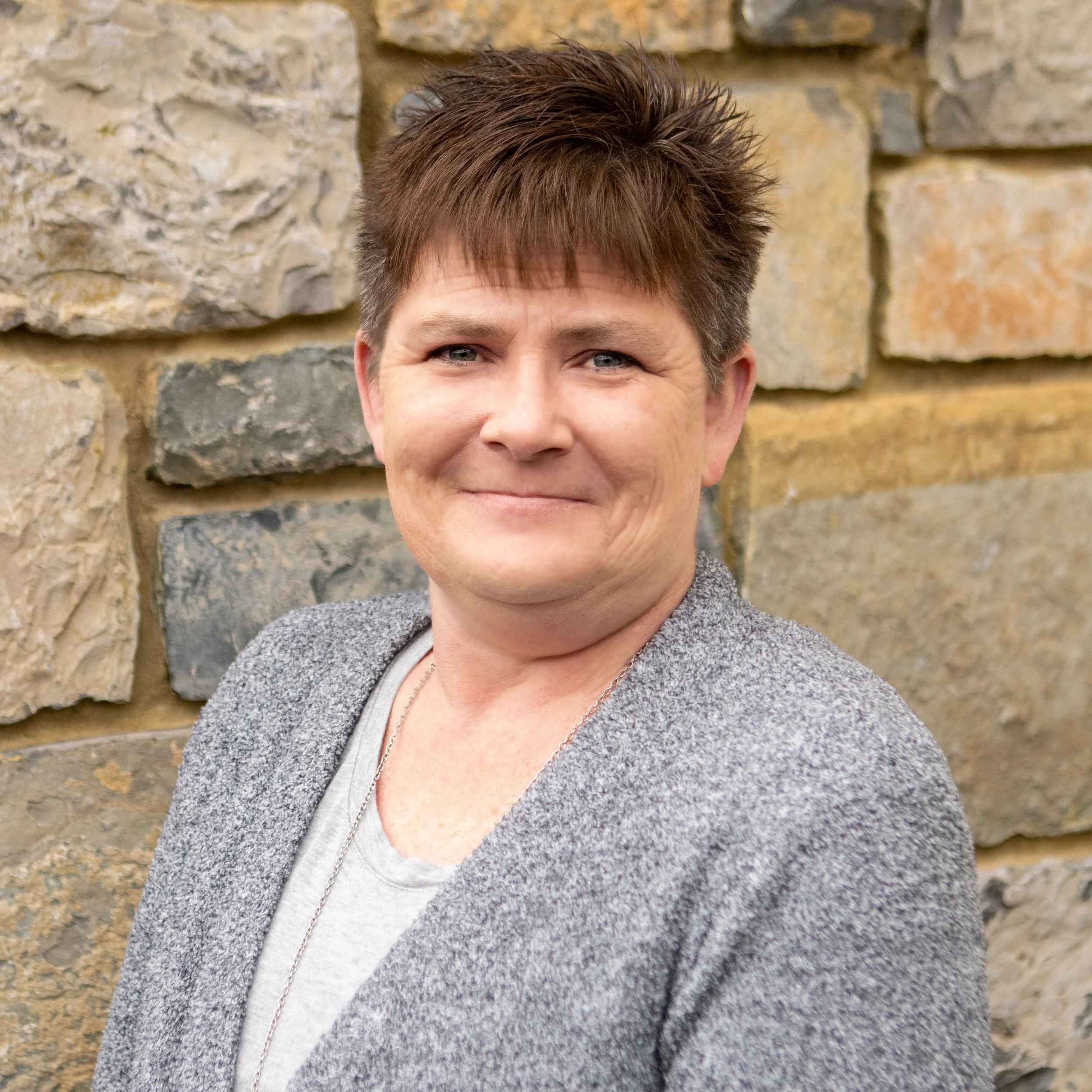 A woman with short brown hair, wearing a gray cardigan and a light gray shirt, standing in front of a stone wall, smiling at the camera.