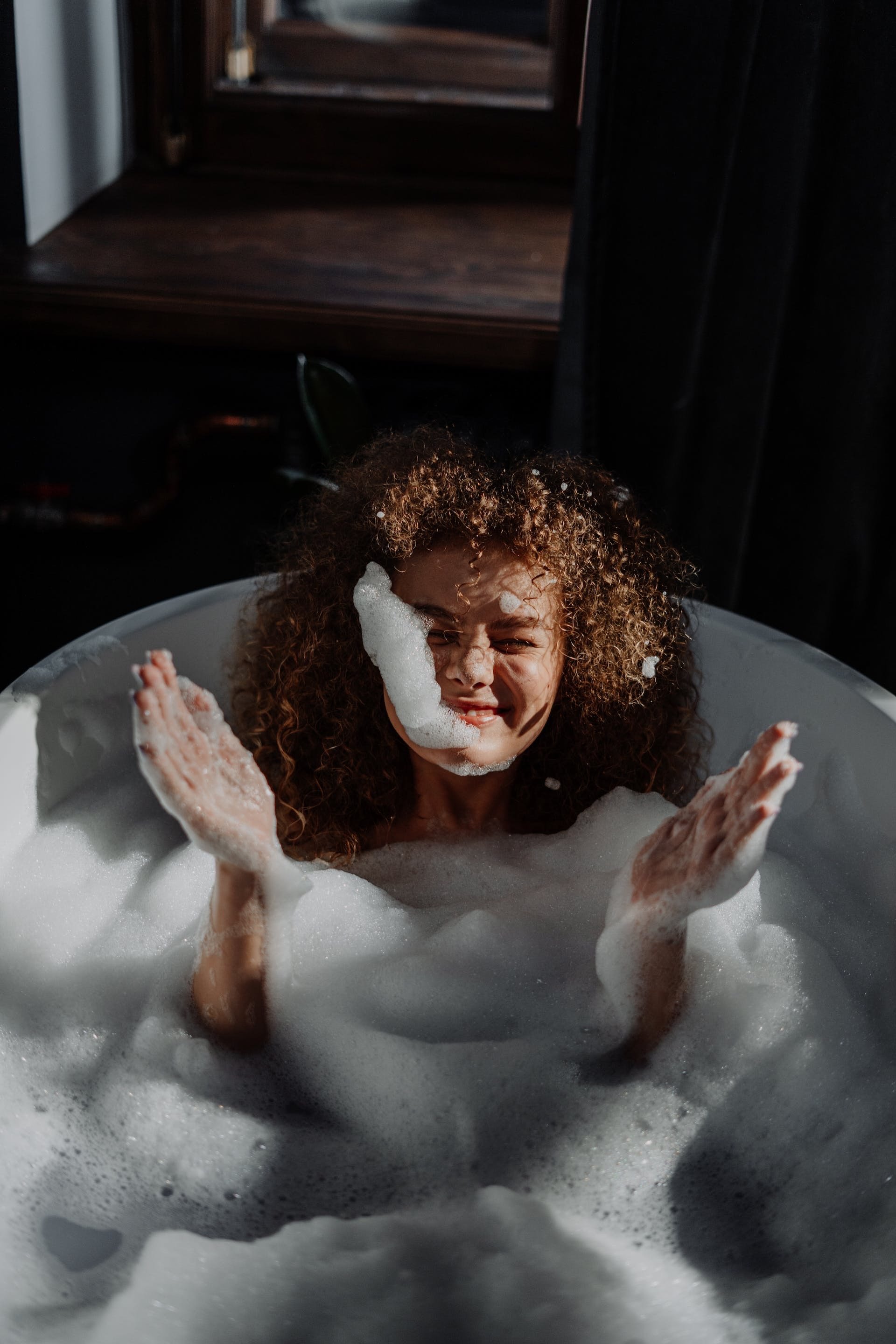 A woman laying in a bubble bath playing with the bubbles and smiling.
